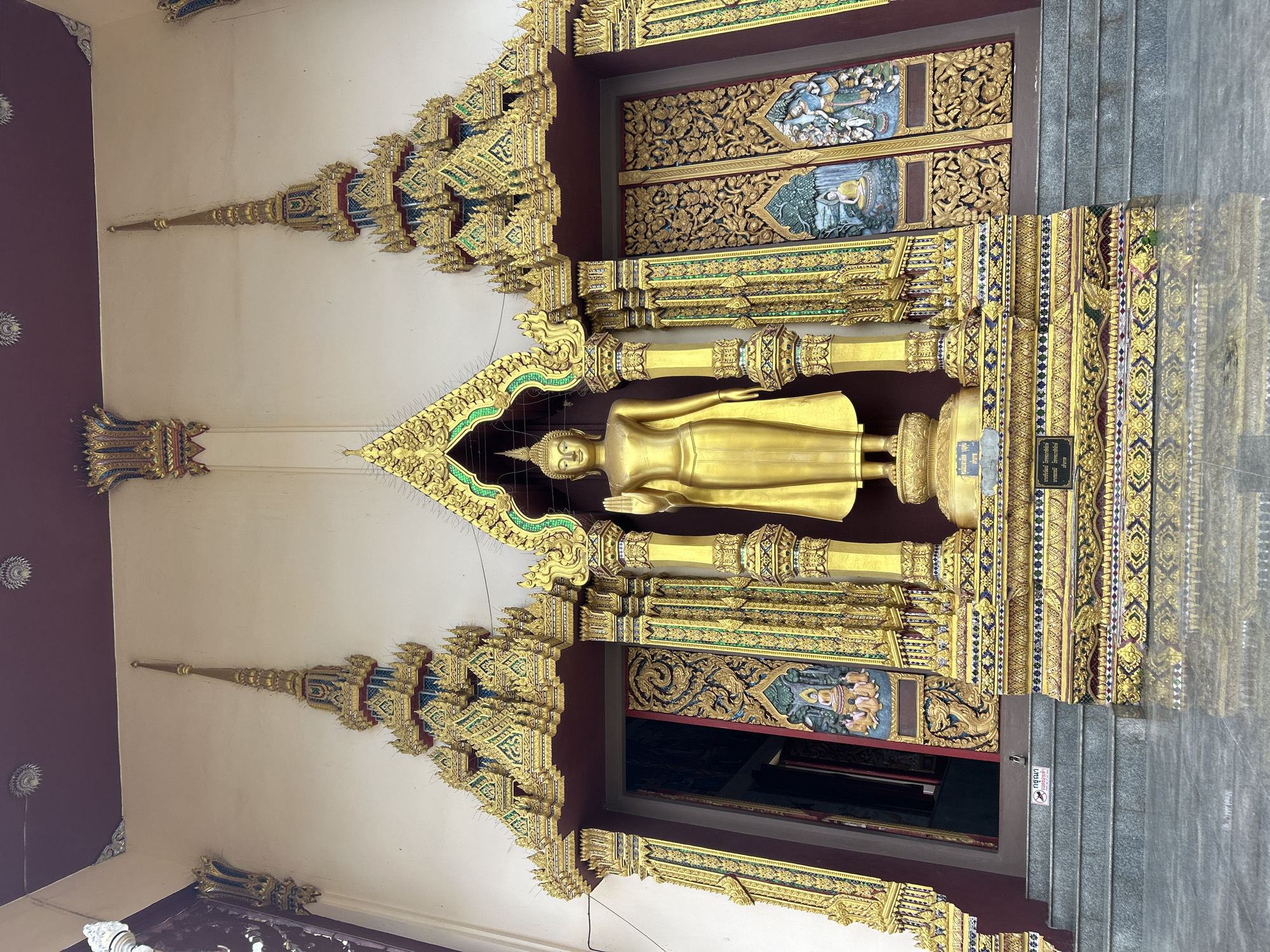 The image shows a large, ornate golden Buddha statue standing on a decorated platform in front of a temple in Ko Samui District, Thailand. The statue is life-sized and depicts the Buddha in a standing posture with his right hand raised. The temple entrance is flanked by two elaborately decorated doorways, featuring golden carvings and intricate patterns. The architecture is typical of Thai temples, with steep, pointed roofs and colorful embellishments. The ground in front of the temple is paved with grey tiles that reflect the golden statue and the surrounding structures. There are steps leading up to the temple entrance. A small sign with text is visible on the steps. The overall impression is one of grandeur and spiritual significance.