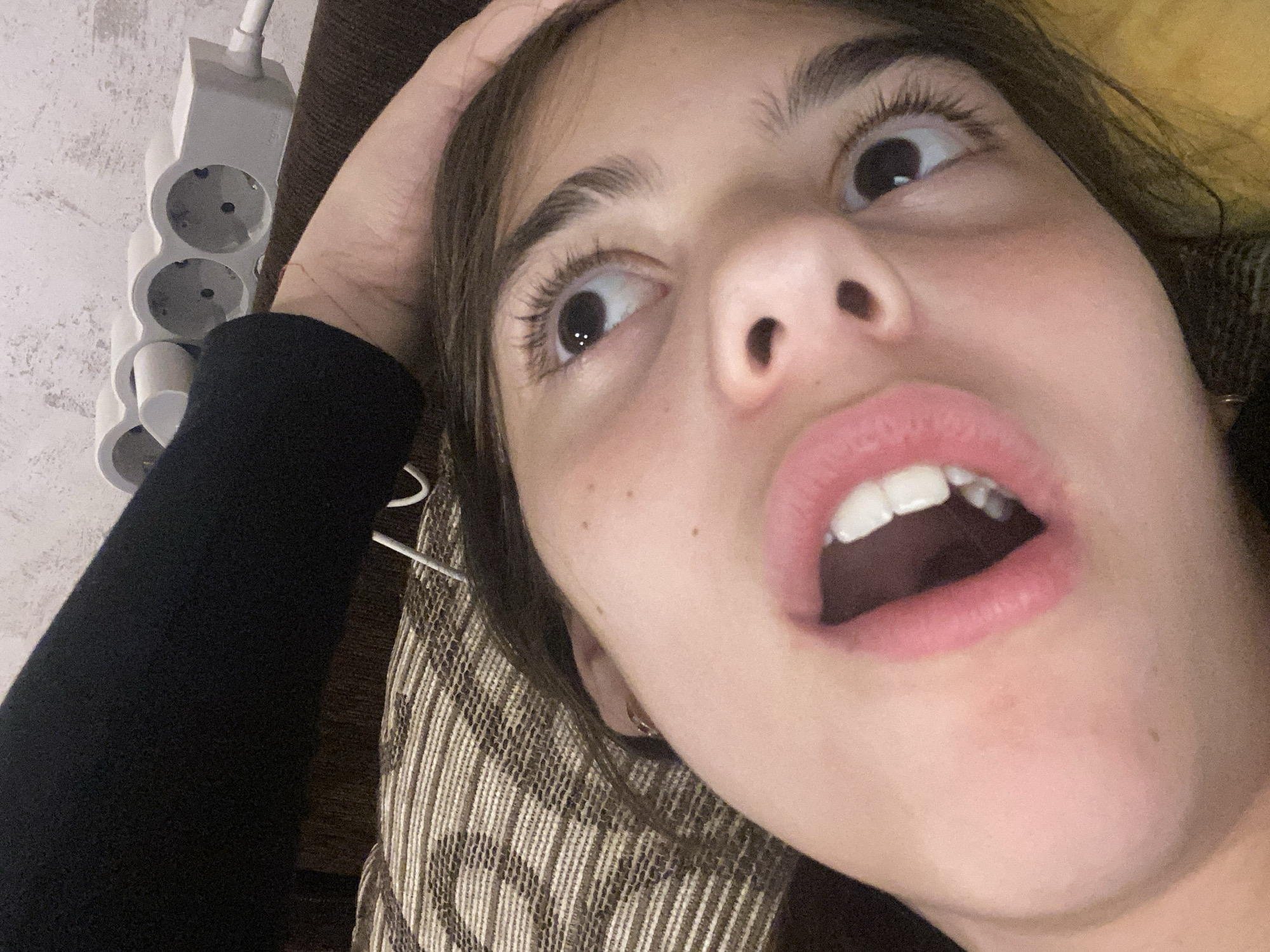 A close-up, low-angle selfie of a young woman with fair skin, dark hair, and brown eyes. Her mouth is open, revealing her upper teeth. Her eyes are wide and looking upwards. She is lying down with her head resting on a patterned pillow or cushion. Her left hand is touching her forehead. A white power strip with European-style outlets is visible in the upper left corner of the image. The background wall is textured and off-white. The location is not identifiable.