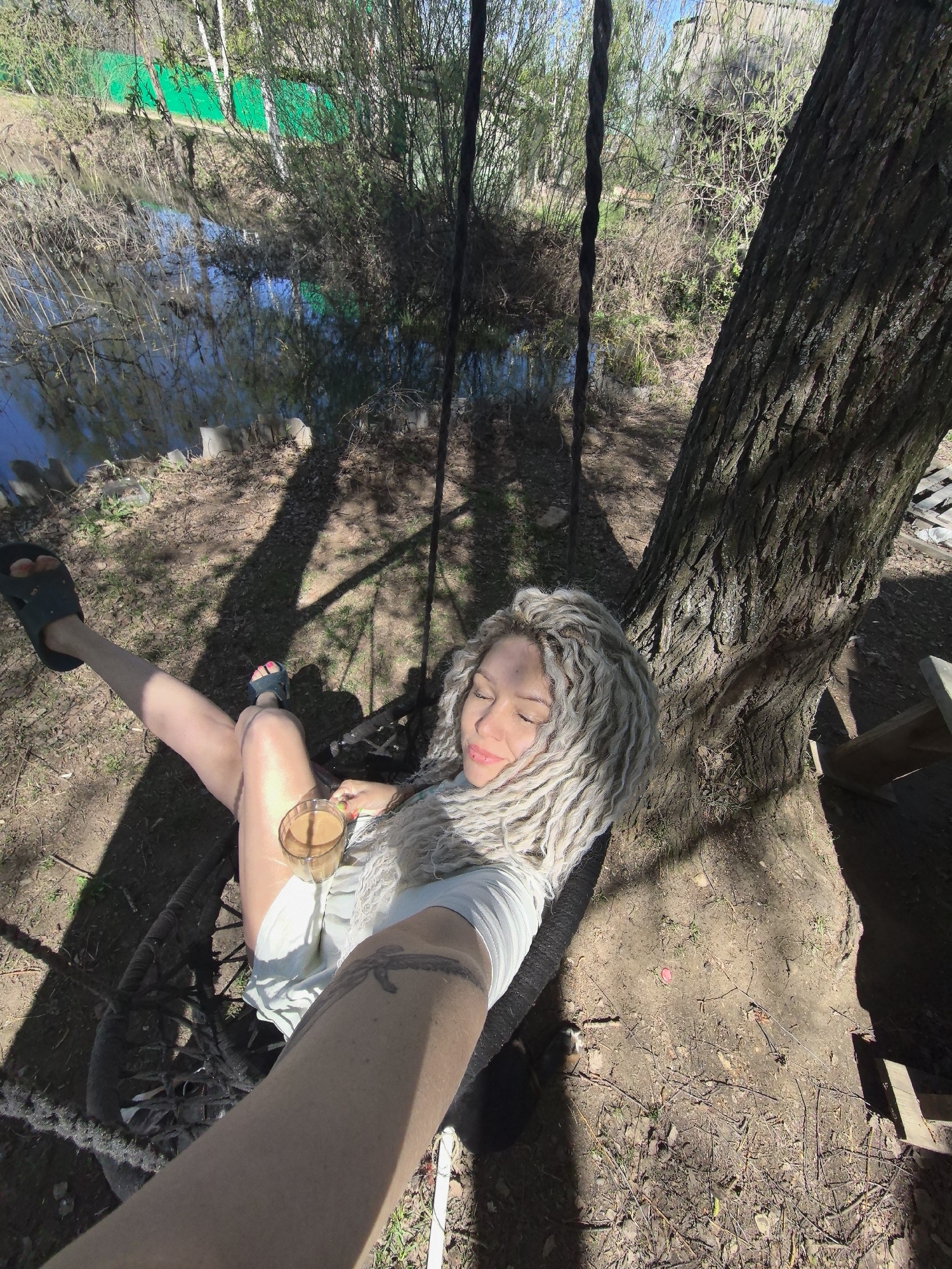 A young woman with bleached dreadlocks is relaxing in a rope swing tied to a tree, seemingly outdoors in a park-like setting in Noviy, Russia. She is wearing a white shirt or dress and has her legs outstretched; one leg is elevated in a sandal. She is holding a glass of what appears to be coffee. A tattoo is visible on her forearm. The ground is covered in dirt and sparse vegetation. Behind her, there is a body of water with reflections. The area is shaded by trees.