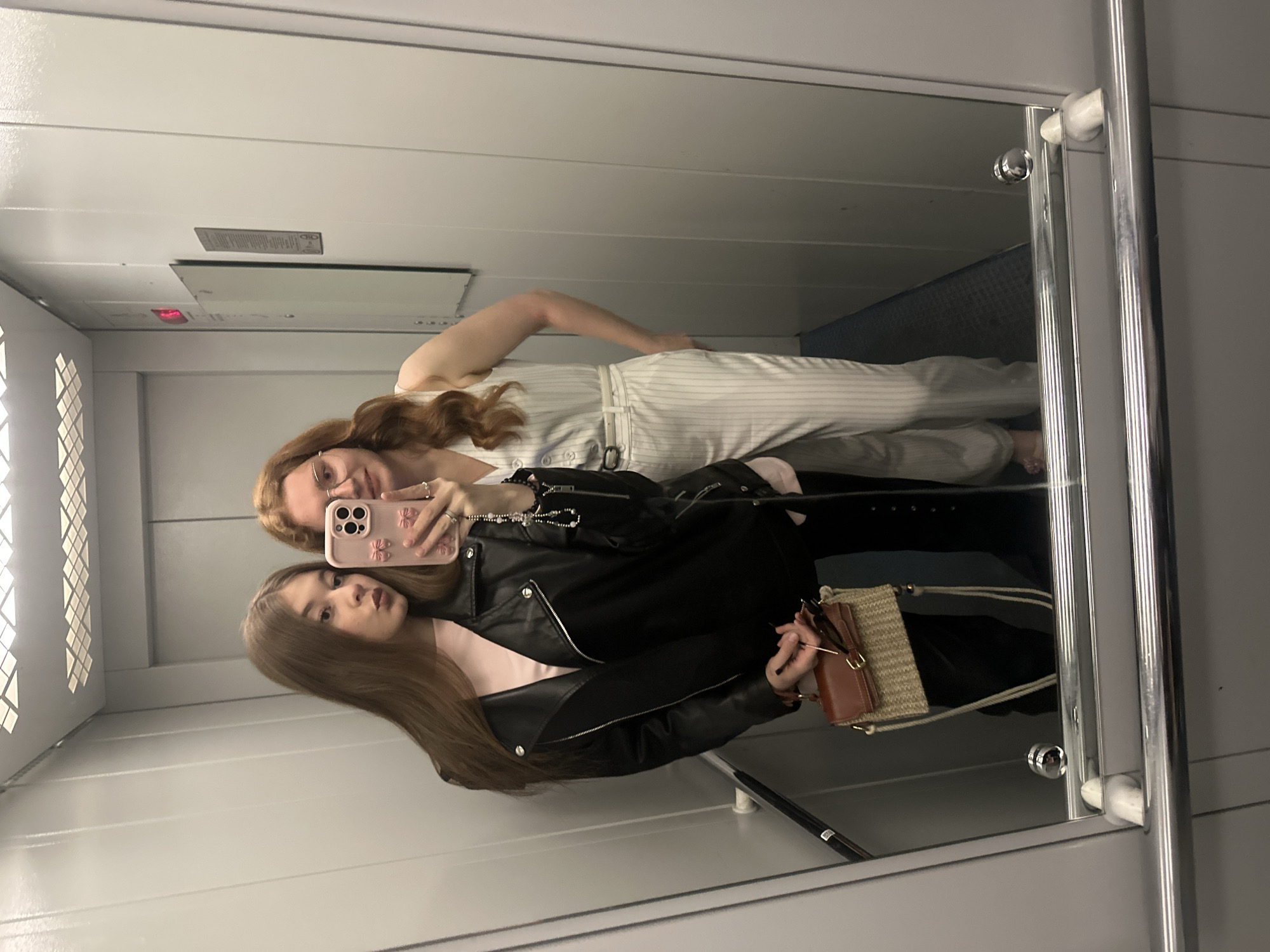 The image displays a mirror selfie of two young women taken inside an elevator, located in Kazan, Russia.
On the left side of the reflection, a woman with long, straight brown hair is visible. She wears a black leather jacket over a light pink top and holds a pink smartphone, adorned with a floral or butterfly pattern, through which the photo is captured. She also carries a small woven bag with a brown leather flap and light-colored straps. Her expression is a subtle pout.
Beside her, the reflection of a second woman with long, wavy reddish-brown hair and eyeglasses is seen. She is dressed in a white pinstriped jumpsuit worn over black pants, and her right hand rests casually on her hip. She has a faint smile on her face. Both women wear rings on their fingers.
The elevator interior is characterized by light grey walls and metallic vertical bars/handrails along the mirror's edge. The ceiling features a long light panel and ventilation grates. On the elevator's back wall, a control panel is visible with a red digital display showing the number '5', likely indicating the current floor. Below this display, a small white rectangular label with some indistinguishable text, including the partially visible letters "AD", and symbols is affixed. The floor of the elevator appears to be a dark blue color, visible in the reflection. The scene is brightly lit by overhead lighting.
