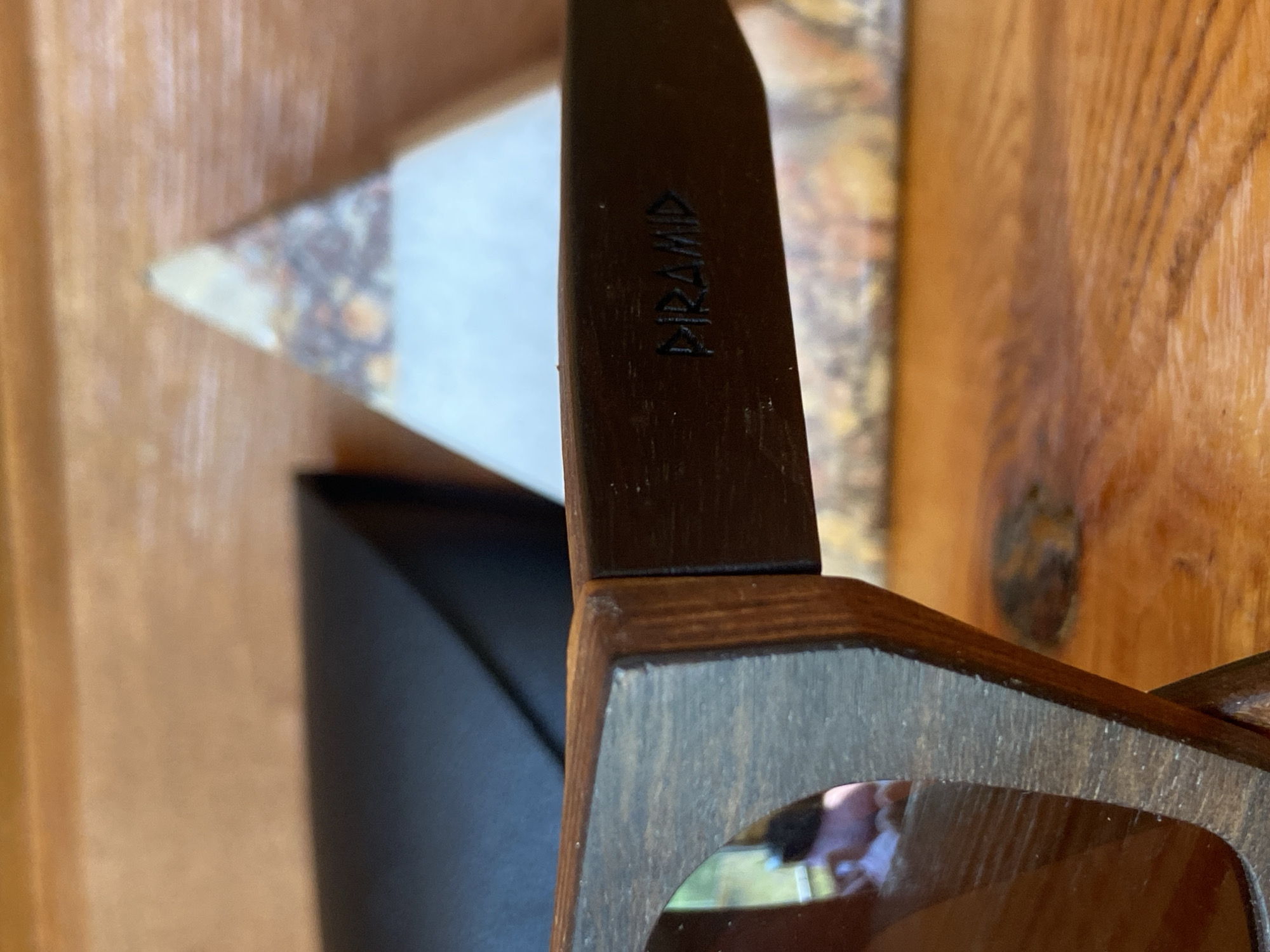 The image shows a close-up of a pair of wooden sunglasses. The sunglasses have dark brown wooden frames and tinted lenses. A logo, which appears to read "PIRAMID", is embossed on the right temple of the sunglasses. The sunglasses are resting on a wooden surface, possibly a table. In the background, there is a black case, and a triangular object with a textured surface. The lighting suggests an indoor setting, with natural light coming from the left. There is no indication of the location being in Красномакское сельское поселение, Ukraine.