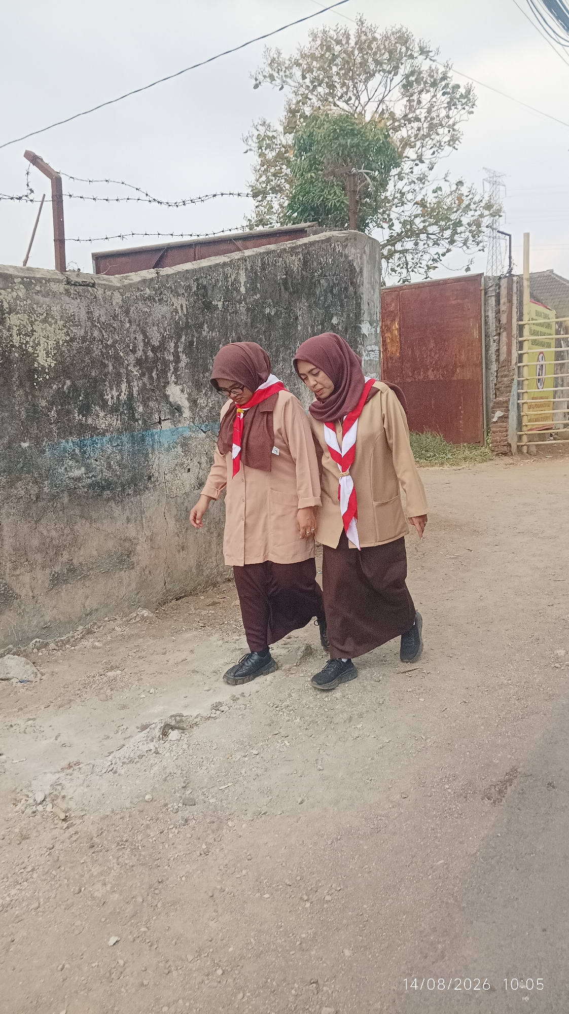 Two women, dressed in tan scout uniforms with maroon hijabs and red and white scout neckerchiefs, walk along a dirt path in Tasikmalaya, Indonesia. The woman on the left wears glasses. The path is lined with a weathered concrete wall on the left and a rusty metal gate and fence on the right. Barbed wire is strung along the top of the wall. In the background, a large tree with sparse foliage stands against an overcast sky, with a transmission tower visible in the far distance. The timestamp "14/08/2026 10:05" is visible in the lower right corner of the image.