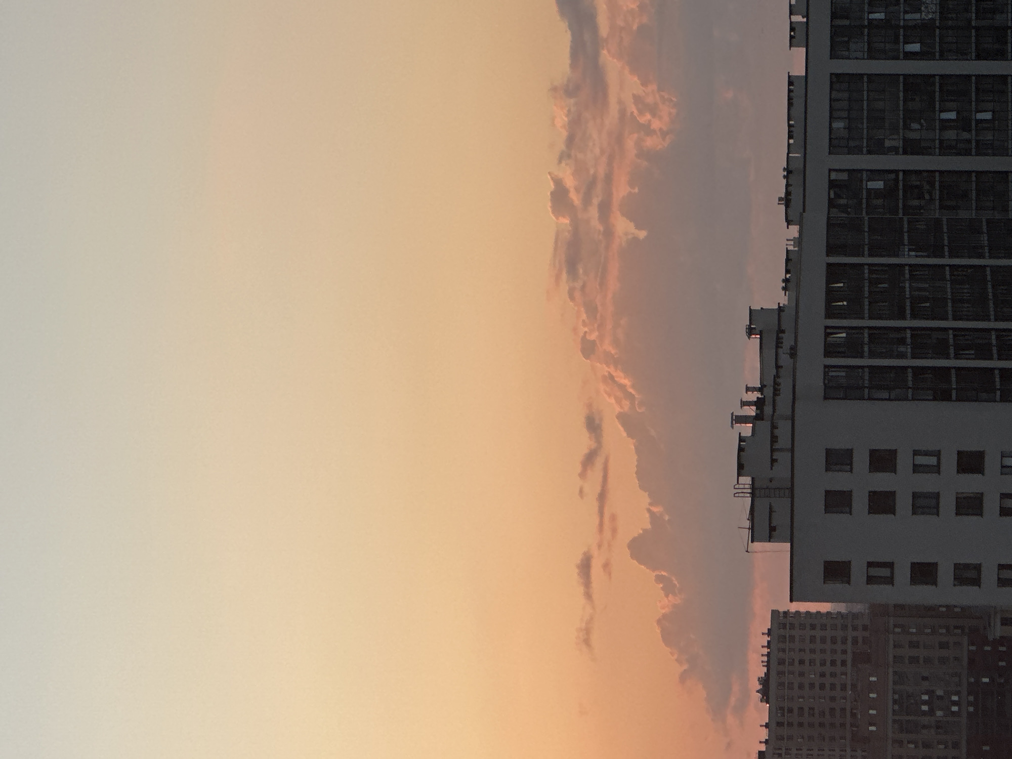 The image shows a cityscape with a sunset sky. The sky is a gradient of warm colors, transitioning from a pale yellow at the top to a peachy orange at the bottom. Wispy clouds are visible, tinged with pink and purple from the setting sun.
Dominating the right side of the frame are several tall buildings. The closest building is a modern structure with a light gray facade and numerous dark, rectangular windows arranged in a grid pattern. Several air conditioning units or similar protrusions are visible along the edge of this building. Further down and slightly behind, another, older building is visible with a darker facade and a denser pattern of windows.
The orientation of the image is such that the buildings appear to be on the left and the sky on the right, suggesting the original photo was taken vertically and then rotated. The scene depicts an urban environment during a sunset. The time of day is clearly indicated by the warm, low-angled light and the colors of the sky. The weather appears clear aside from the clouds. No people are visible, and there is no visible text in the image.
