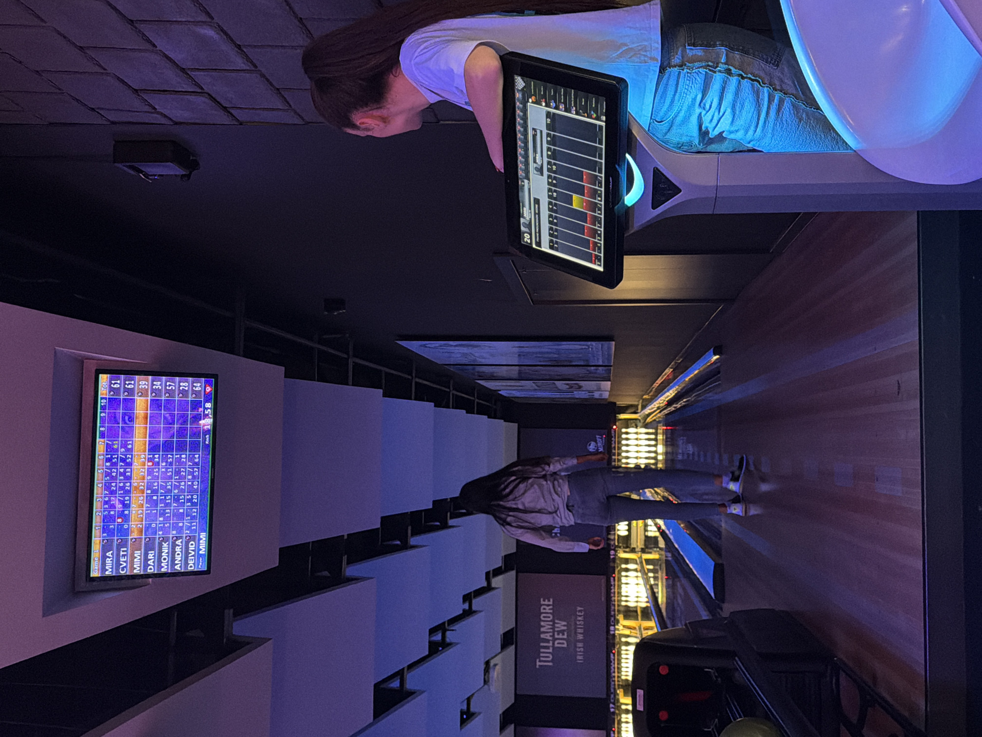 The image depicts the interior of a modern bowling alley in Sofia, Bulgaria, characterized by dim ambient lighting with purple and blue LED accents. The scene is viewed from behind one of the bowling lanes, looking towards the pin decks.
On the far right, a wall features a dark, textured surface resembling stacked stone or large tiles. The ceiling above is dark, with angular, suspended white acoustic panels illuminated from behind. In the distance, multiple bowling lanes are visible, brightly lit along their length, leading to illuminated bowling pins at the far end. A large screen in the background displays "TULLAMORE DEW IRISH WHISKEY."
Above the lanes, a large scoreboard screen displays the current game scores for multiple players: MIRA (61), CVETI (61), MIMI (39), DARI (34), MONIK (57), ANDRA (28), and DEIVID (64). Player MIMI is currently active.
In the immediate foreground on the right, a person, identified as female by her long, dark brown hair tied in a high ponytail, stands leaning with her right arm on a sleek, grey-white interactive touchscreen control panel. She is wearing a white short-sleeved t-shirt and light blue jeans, with her back mostly to the camera, observing the lanes. The control panel emits a blue glow from its base and displays a detailed score and frame progression.
To the left, on one of the bowling lanes, another person, also appearing female based on build and long, dark hair, is walking away from the camera. She wears a light-colored long-sleeved shirt and blue jeans, with sneakers featuring white soles and dark uppers. Her action suggests movement on the bowling approach. A dark bowling ball return mechanism with a yellow bowling ball is partially visible on the far left foreground. The overall environment indicates an active bowling game in progress.