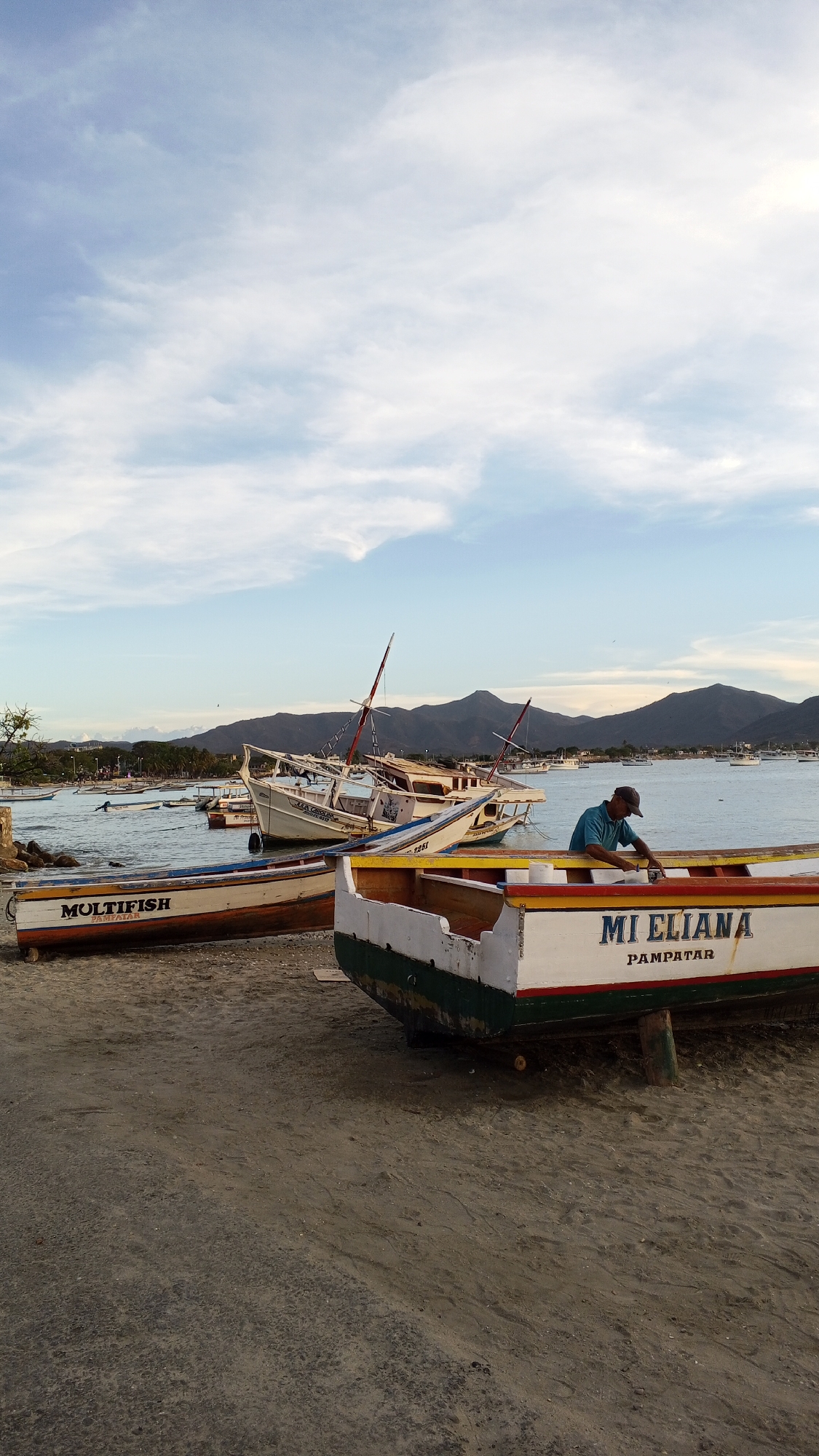 The image shows a coastal scene in Pampatar, Venezuela with several boats on a sandy beach, likely used for fishing. In the foreground, there are two boats labeled "MULTIFISH" and "MI ELIANA PAMPATAR" on the beach. A man wearing a blue shirt and a cap is working on the "MI ELIANA" boat. In the background, other boats are in the water near the shoreline with mountains visible in the distance under a partly cloudy sky. The environment appears to be a fishing village or harbor.