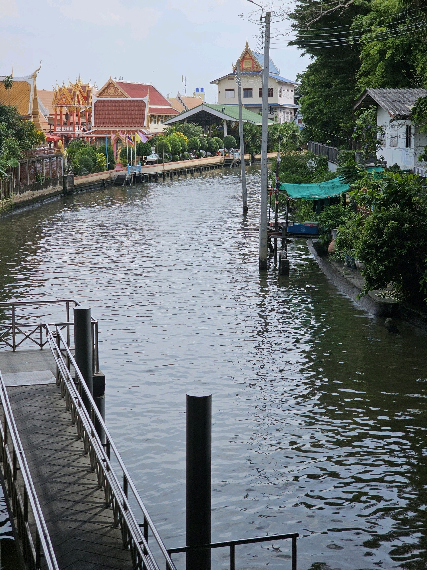 The image captures a canal in Bangkok, Thailand, with buildings and lush greenery lining its banks. A metal ramp with handrails leads down to the water's edge on the left side of the frame. The canal itself is wide and calm, with gentle ripples reflecting the overcast sky.
On the right bank, dense foliage obscures some of the structures. A small white building with a gray tiled roof is visible, partially covered by vines. Further down the bank, a section of a temple complex is apparent, characterized by its ornate, tiered roofs and vibrant colors. The temple features golden spires and intricate detailing, typical of Thai architecture. In front of the temple, flags are displayed, and a walkway or pier extends into the water.
On the opposite bank, another temple structure with a red tiled roof and decorative gable is prominent. Other buildings, possibly residential or commercial, are nestled behind the temple. A utility pole stands in the middle of the canal, its wires stretching across the water. A green tarp is draped over a structure near the right bank, possibly an awning or shelter. The overall scene suggests a tranquil, yet inhabited, urban waterway, blending natural elements with architectural structures.