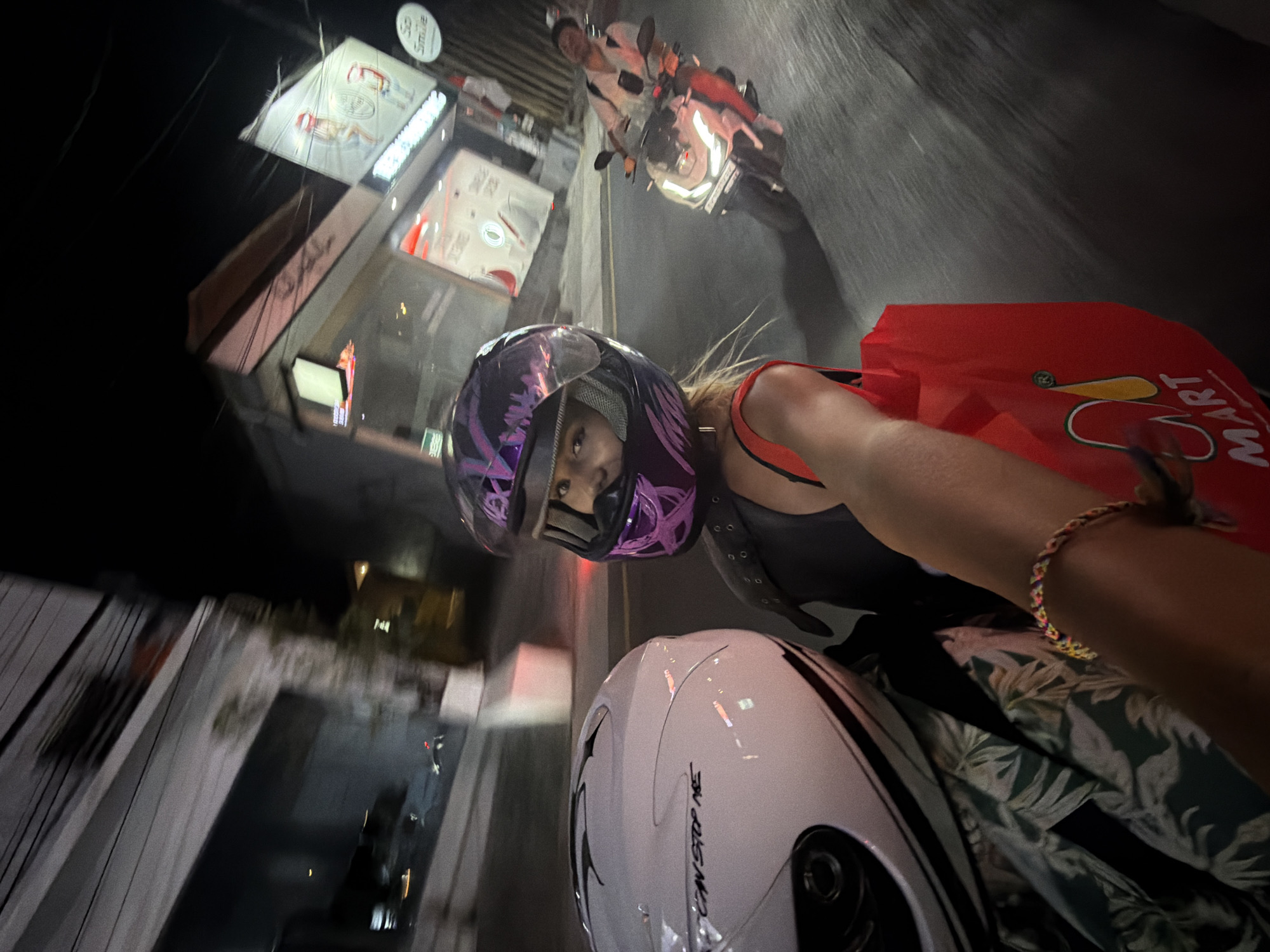 The image captures a first-person perspective of a person riding on the back of a scooter at night in North Kuta, Indonesia. The rider in the foreground is wearing a purple and black helmet with reflective purple accents, a black top, and a red bag is visible, held by their hand. To the left, a white helmet is also visible, suggesting the rider is a passenger. In the background, another person is riding a white scooter. The scene appears to be on a street lined with buildings, some of which have illuminated signs, indicating a commercial area. The motion blur suggests the scooters are in motion. The lighting indicates it is nighttime.