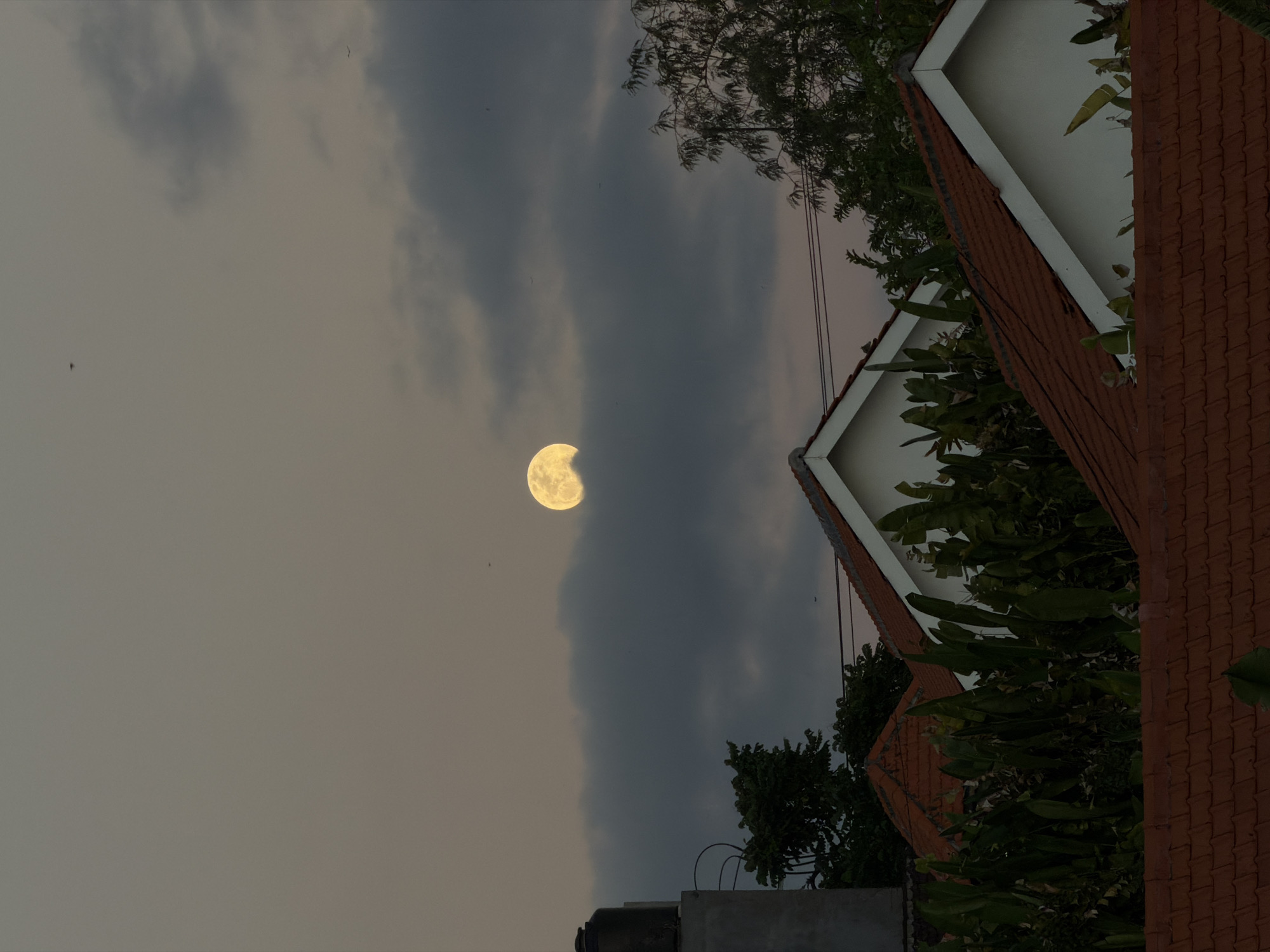 This image captures a scene in North Kuta, Indonesia, featuring a full moon partially obscured by dark, wispy clouds. The moon appears a luminous yellow, casting a soft glow. In the foreground, terracotta tiled rooftops of houses are visible, interspersed with lush green foliage. The architecture suggests a residential area. The sky is a muted grey-brown, indicative of twilight or an overcast day. A dark, cylindrical water tank is perched on a grey structure on the left side of the frame, possibly a rooftop. Several trees with dark green leaves frame the upper right portion of the image. There are no people visible in the scene. The overall atmosphere is serene and quiet.