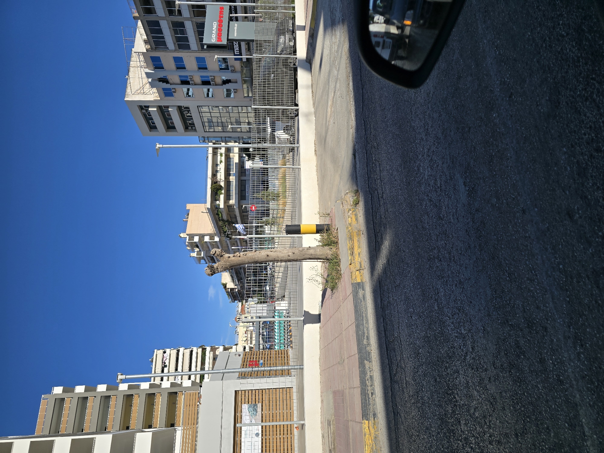 The image depicts a street scene in Athens, Greece, under a clear blue sky. In the foreground, the asphalt road surface is visible, with a car's wing mirror reflecting traffic in the distance. A sidewalk with yellow and gray curbs runs alongside the road. A row of concrete buildings with balconies and windows lines the street. A metal fence encloses a parking area with several cars and shopping carts visible. A trimmed tree stands in front of the fence, next to a black and yellow striped bollard. A sign with Greek text and a red logo that reads "GRAND" is visible on one of the buildings. The overall impression is of a typical urban environment in Greece.