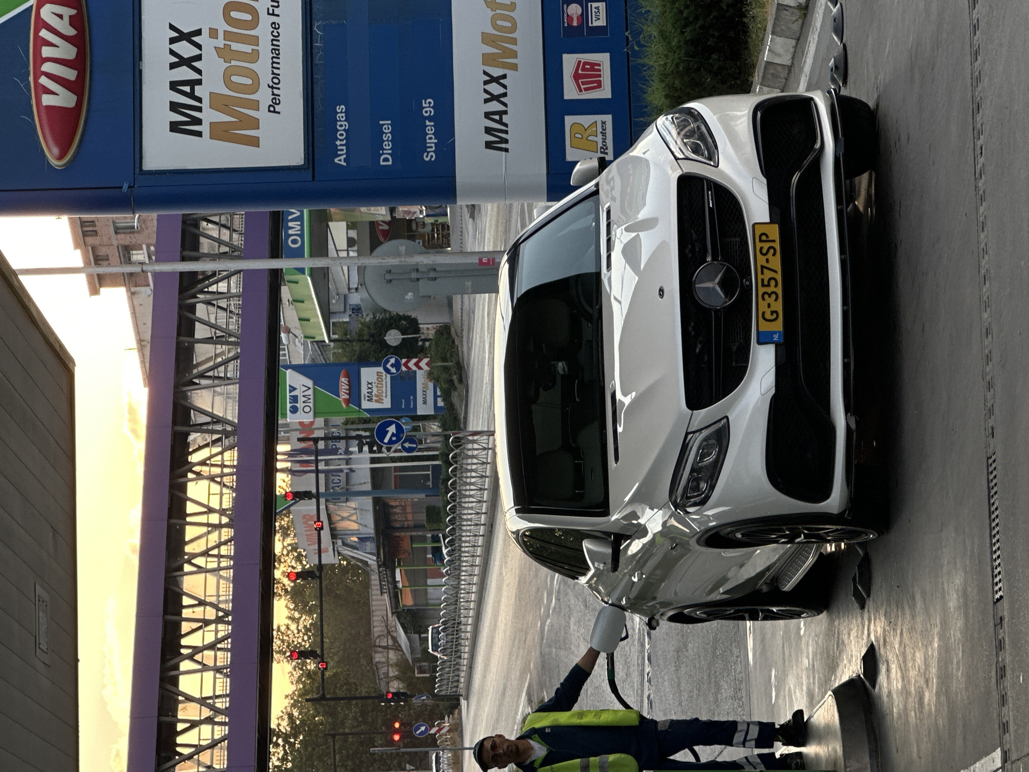 A person wearing a reflective vest and a cap is fueling a white Mercedes-Benz SUV at a gas station. The gas station has signs for "VIVA MAXX Motion" and OMV. The license plate of the car is visible and reads "G-357-SP". The scene appears to be a typical gas station environment with pumps and signage. The lighting suggests it is either early morning or late afternoon, with warm, soft sunlight creating long shadows. The overall atmosphere is neutral, focusing on the task of refueling the vehicle.