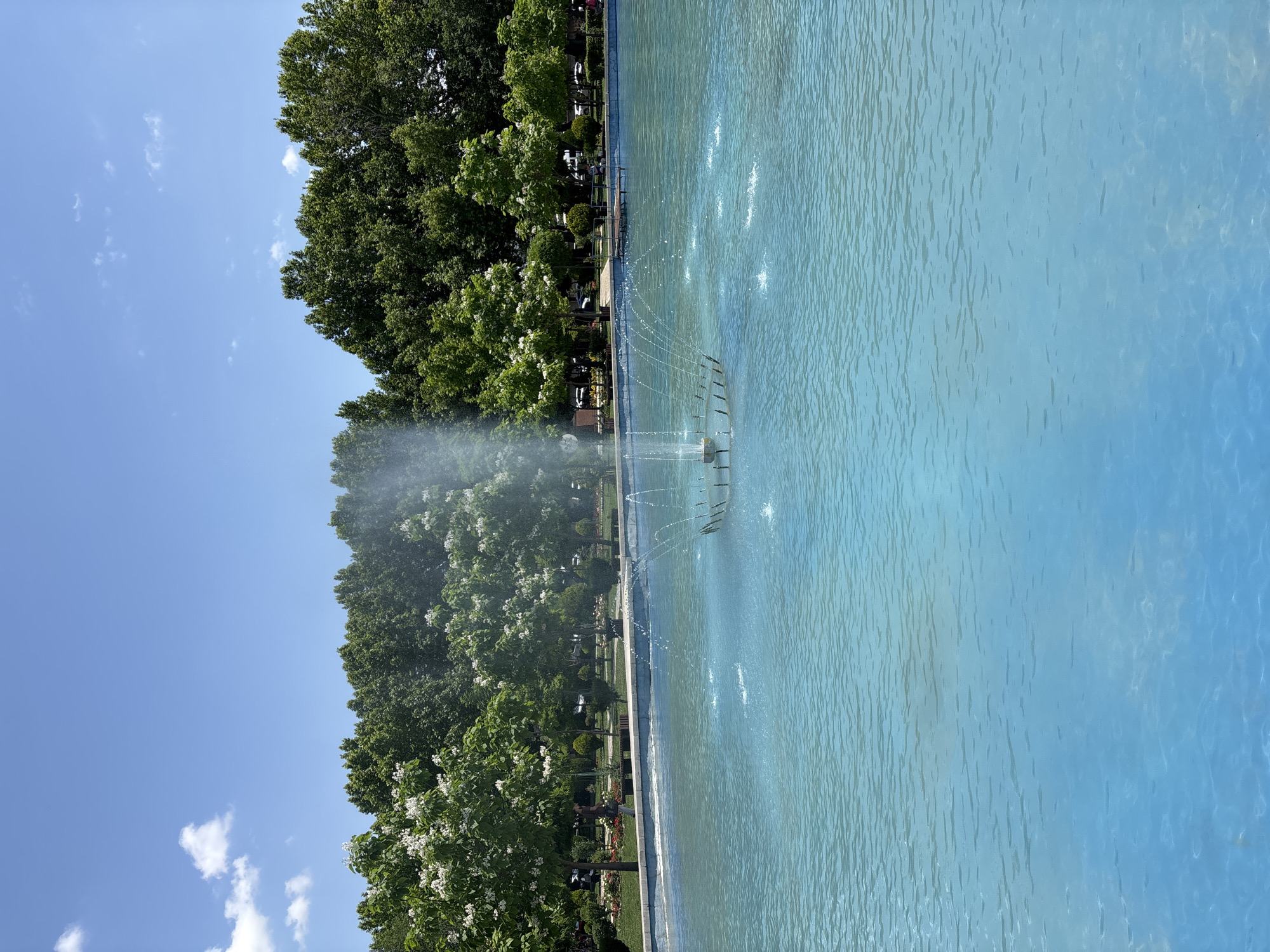 The image captures a serene scene in Yerevan, Armenia, likely within one of its many beautiful parks. A large, bright blue pool occupies the foreground, its surface rippled by gentle breezes and disturbed by the graceful arc of a fountain in its center. This fountain, a circular metallic structure, propels water upwards and outwards in delicate streams, creating a refreshing mist.
Beyond the pool, a manicured park unfolds. Lush green trees, some adorned with white blossoms, form a dense canopy, creating a vibrant backdrop. A few individuals can be seen strolling or relaxing on benches along the park's edge, their presence adding a touch of life to the tranquil setting. The clear blue sky above, dotted with a few wispy clouds, suggests a pleasant day, perfect for enjoying the outdoors. The overall impression is one of peaceful urban greenery, a welcome oasis in the heart of the city.