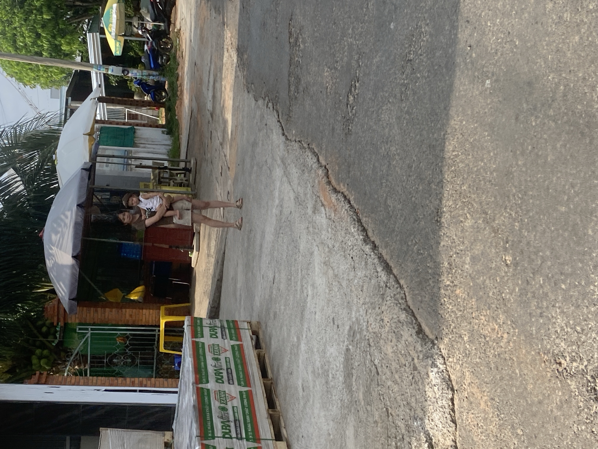 A woman is holding a small child in her arms on a paved street in Mũi Né, Vietnam. The woman is wearing a brown t-shirt and shorts. The child is wearing a white shirt and brown shorts. They are standing near a white umbrella, which is providing shade. In the background, there are buildings, trees, and motorbikes. The street is cracked and worn. To the left of the image, there are stacks of materials wrapped in plastic. The overall impression is of a casual, everyday scene in a warm climate.