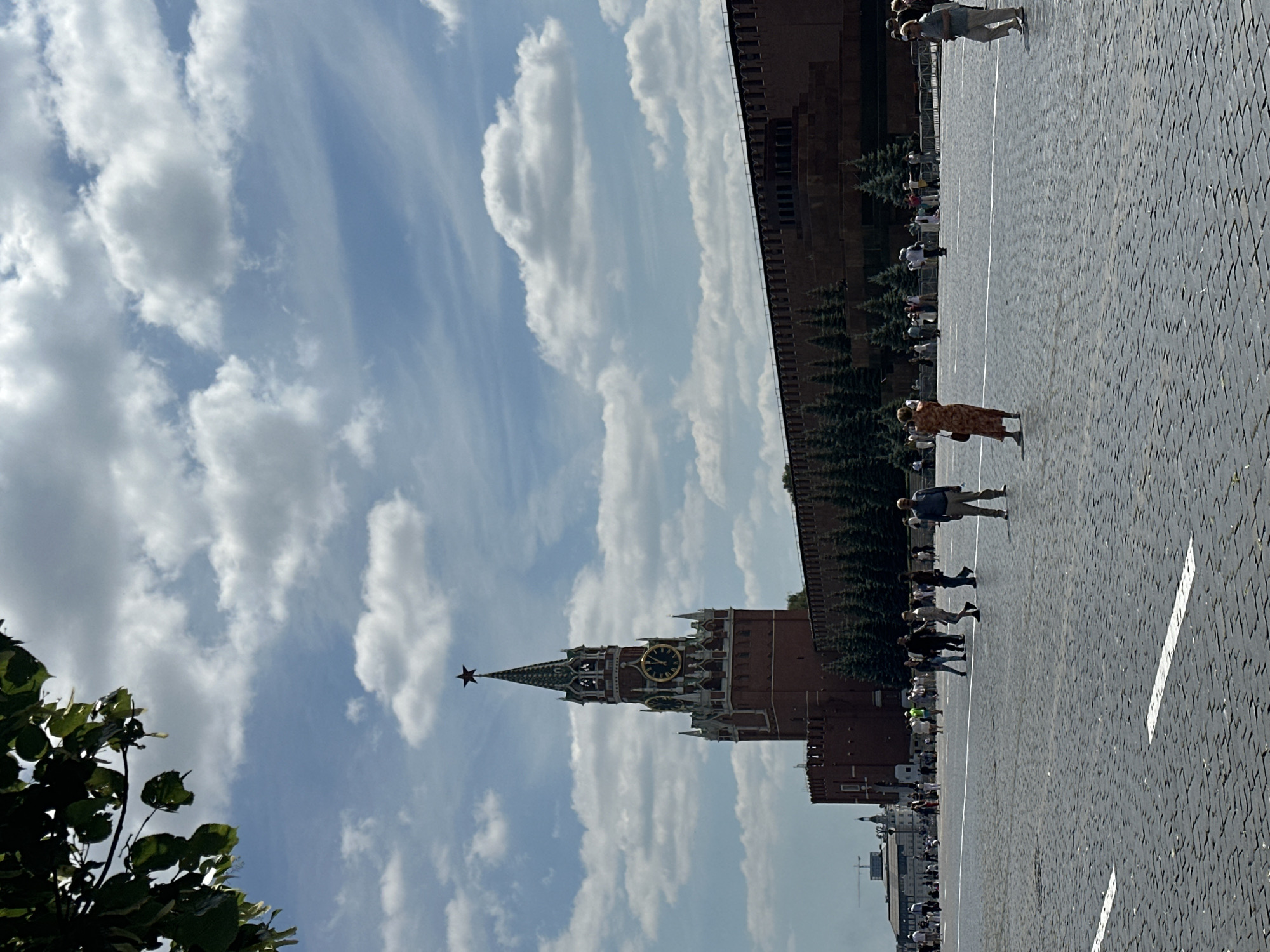 The image depicts Red Square in Moscow, Russia, on a bright, partly cloudy day. In the background, the iconic Spasskaya Tower, part of the Moscow Kremlin, stands tall with its clock face visible and a red star atop its spire. Adjacent to the tower is the Kremlin Wall, a long, red brick structure with crenellations along its top. In front of the wall, a line of dark green fir trees is visible.
The foreground is dominated by the vast, cobblestone expanse of Red Square, marked by white painted lines. Scattered across the square are numerous people. In the middle ground, a woman in a patterned dress is standing, facing away from the camera, and appears to be taking a photograph with her phone. To her right, a man in a light-colored shirt and pants is walking away from the camera. Other individuals and small groups are visible further in the distance, dispersed throughout the square. To the far left, a section of green foliage from a tree hangs into the frame.