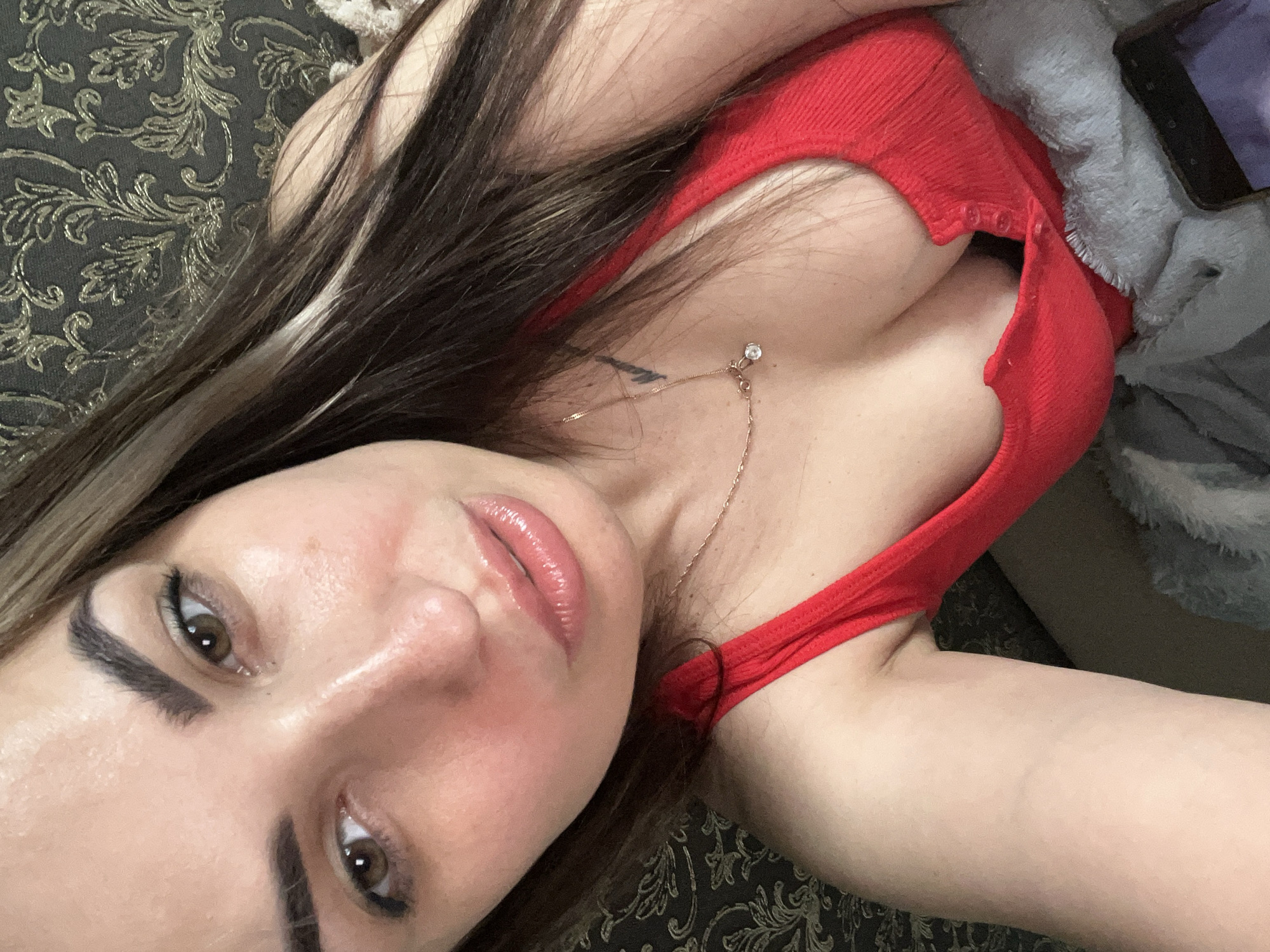 A close-up, high-angle selfie depicts a young adult female with long, dark brown hair featuring lighter brown streaks. Her hair falls over her left shoulder and chest. She possesses brown eyes and full, glossy lips. Her eyebrows are dark and well-defined. Visible on her skin are areas of shine, particularly on her forehead, nose, and cheeks, with a slight reddish tone to the cheeks. A subtle tan line is present across her forehead. She wears a red, ribbed tank top with a partial button-front closure, which is open to reveal cleavage. A delicate gold-colored chain necklace with a small, clear gemstone pendant and a smaller secondary charm is draped around her neck. A small, dark, stylized tattoo is visible on her upper chest/shoulder area to the viewer's right. The background consists of a dark wall or upholstery with an intricate metallic gold floral or damask pattern. A grey, textured blanket or throw is visible in the lower portion of the image. A black smartphone with its screen partially visible lies on the blanket in the lower right corner. The specific location in the city of Unknown, Unknown, cannot be determined from the image; it appears to be an indoor setting.