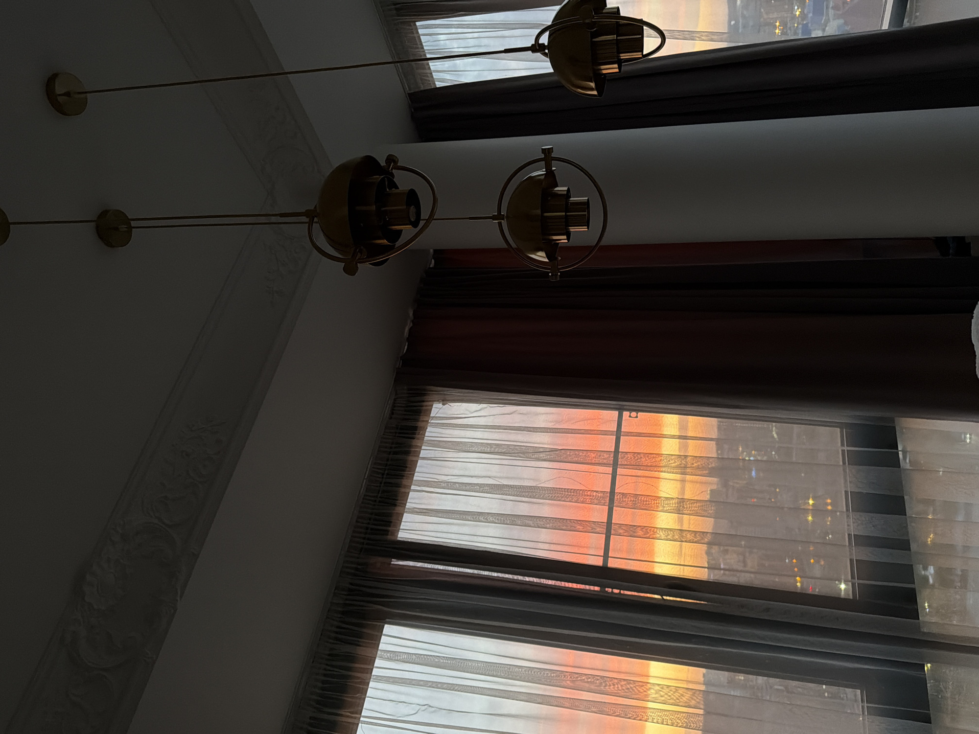 The image depicts an interior scene in what appears to be a residential building, likely an apartment, with a partial view of a cityscape seen through a window. The illumination suggests it is either sunrise or sunset.
**Objects and Environment:**
* **Window:** Large windows are a dominant feature, partially covered by light-colored sheer curtains and darker drapes.
* **Cityscape:** The window offers a view of a city with buildings and lights, likely Krasnoyarsk, Russia given context.
* **Light Fixture:** A modern, multi-bulb, gold-colored chandelier hangs from the ceiling.
* **Ceiling:** The ceiling has decorative molding.
* **Color Palette:** The scene is characterized by soft light, warm tones from the sunrise/sunset, and neutral colors from the walls and curtains.
**Action and Interaction:**
* The light from the exterior is interacting with the interior, creating a warm ambiance.
* There are no explicit actions or interactions taking place between people in the image.
**Note:** The description is based solely on the provided image and assumes the location is Krasnoyarsk, Russia. More information would be needed for further detail.