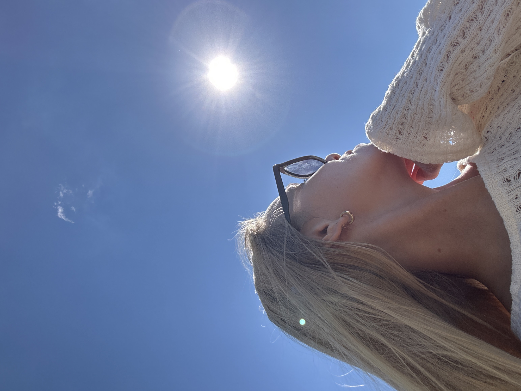 A woman with blonde hair and sunglasses is looking up at the sun. She is wearing a white sweater. The sun is shining brightly in a clear blue sky with a few wispy clouds. The image appears to be taken outdoors, possibly in a park or open area, given the clear sky and sunlight. The location of Gornyi Erik, Russia is not discernible from the image content.