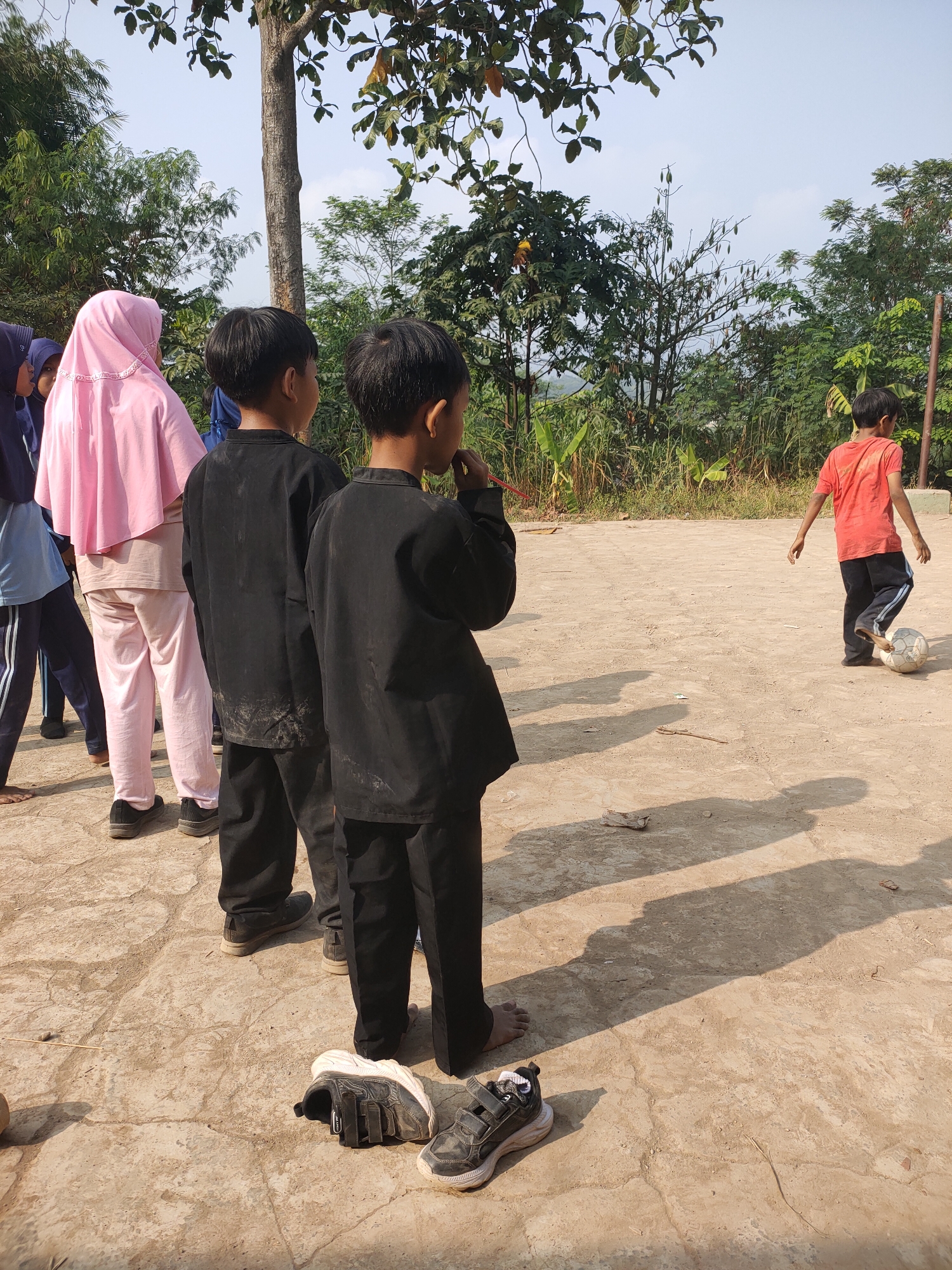 In a dusty, outdoor field, likely in Subang, Indonesia, several children are engaged in a casual game of soccer. Two boys, dressed in matching black uniforms, stand facing away from the camera, their attention fixed on a third boy in an orange shirt who is actively kicking a soccer ball. The boy in orange, wearing dark pants with a blue stripe, appears to be mid-kick. In the background, a woman in a pink hijab and light pink pants stands with other children, observing the game. The environment is characterized by dry, cracked earth, sparse vegetation, and trees. Sunlight casts distinct shadows on the ground, indicating it is a bright, sunny day. The presence of fallen leaves and the overall arid appearance suggest a dry season. Two pairs of athletic shoes lie on the ground in the foreground, indicating that at least some of the children have removed their footwear to play.