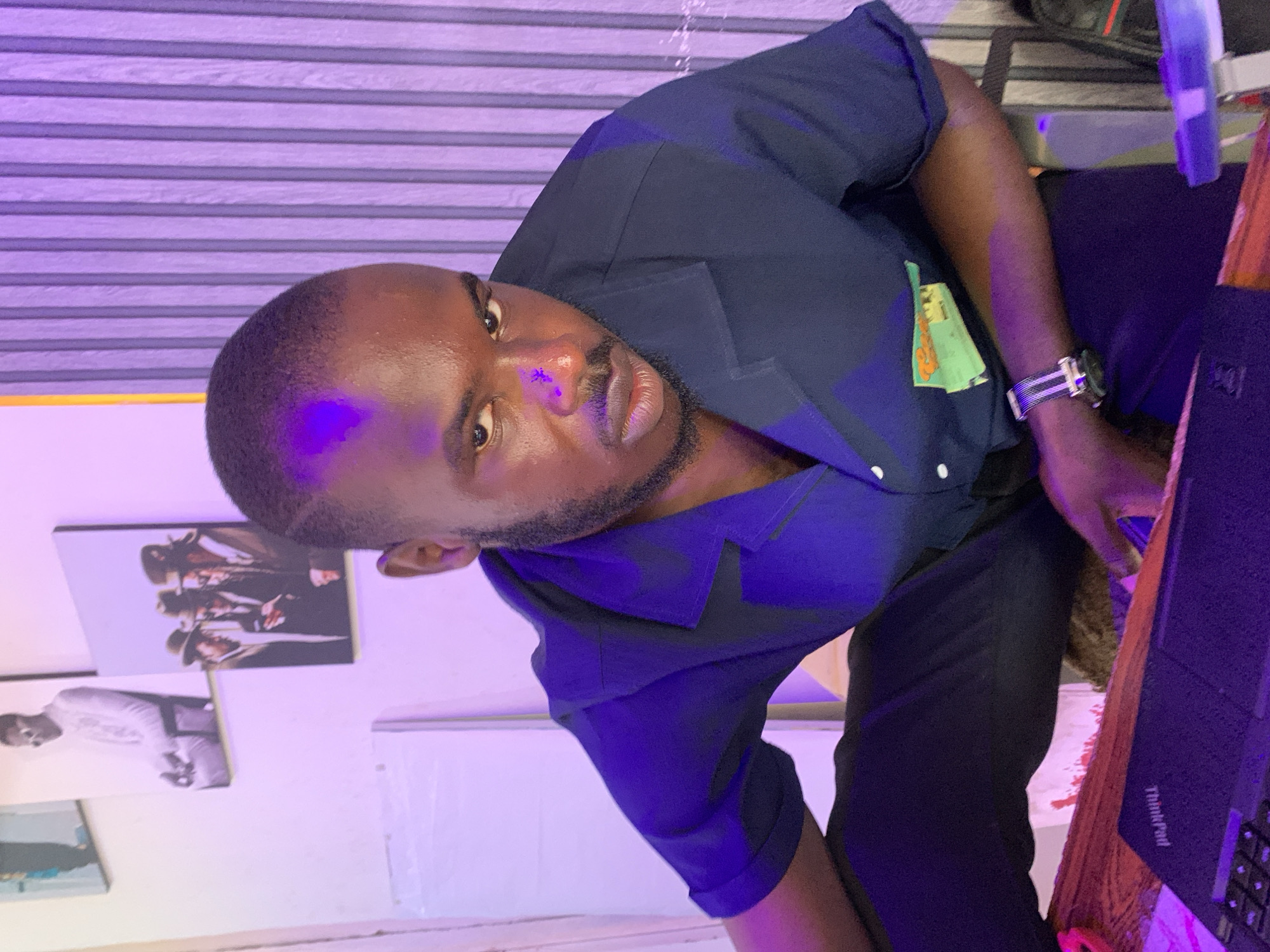 A man of African descent with a short haircut and a beard is captured in a close-up, downward-angled shot. He wears a dark blue collared shirt with the sleeves rolled up, revealing a watch on his left wrist. His gaze is directed towards the camera with a serious expression. The man is seated in what appears to be an indoor setting, indicated by the wall with vertical panels in the background, which is illuminated with purple lighting. On the wall, there are three framed pictures. The one on the far left is a black and white image of a person in a standing pose. The middle picture is also in black and white, showing a person in a seated position with sunglasses. The picture on the right displays three individuals wearing hats and suits. In the foreground, a laptop with the "ThinkPad" logo visible is placed on a wooden surface. The overall lighting is dim, with a strong purple hue dominating the scene, suggesting ambient or artificial lighting. The scene is not specifically identifiable as being in Jos, Nigeria, as there are no explicit visual cues to confirm its location within that city.