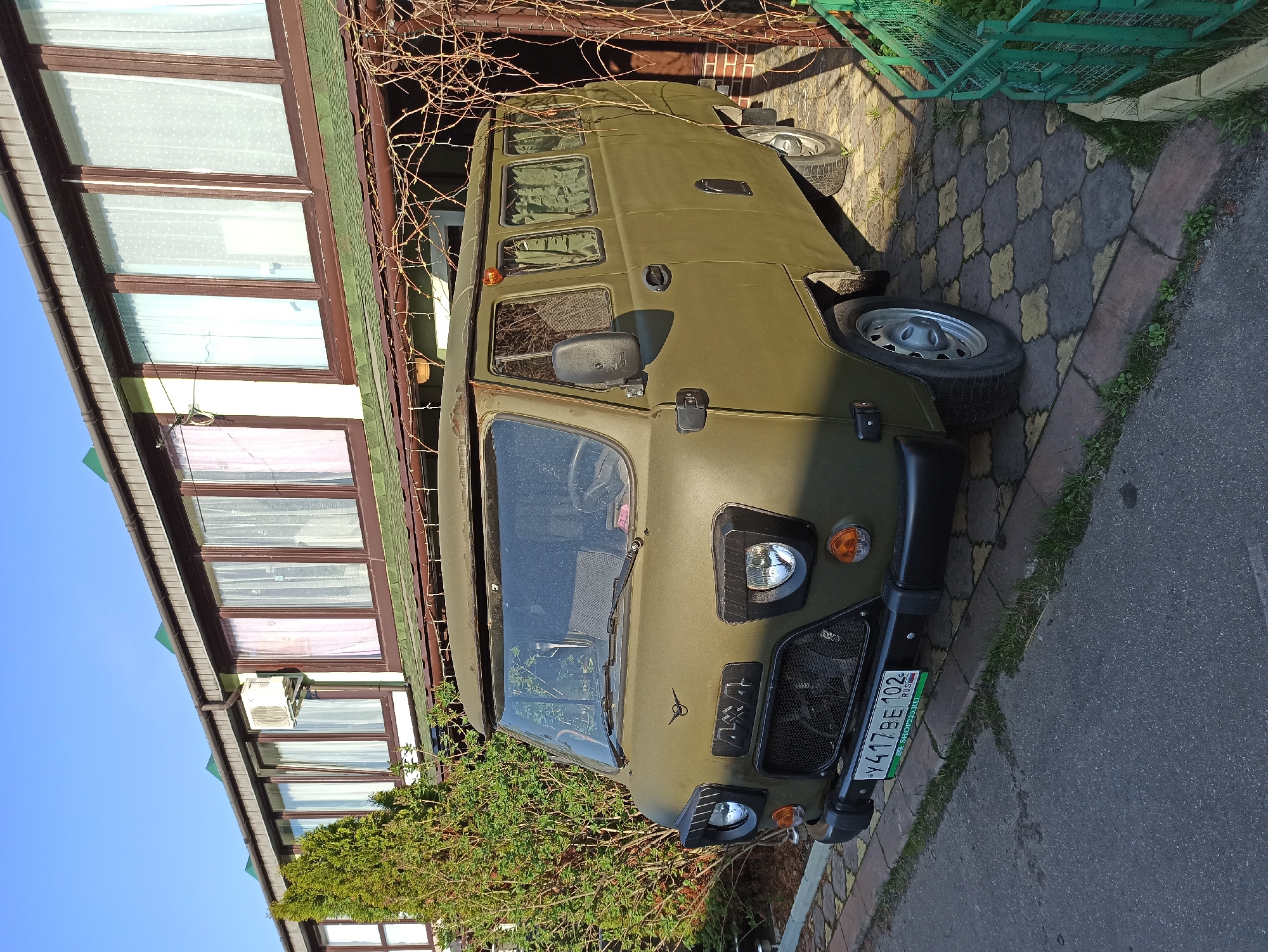 The image shows a parked, olive green UAZ-452 van. The vehicle is parked on a brick and asphalt surface, adjacent to a building with a multi-windowed facade, likely a commercial or residential structure. The building's design features several windows with white curtains and brown frames. A small section of green metal fencing is visible to the right of the van. The license plate is visible on the front bumper and indicates the location as Рыбхоз, Russia. The scene is captured during daylight under a clear sky. No people are visible in the frame.