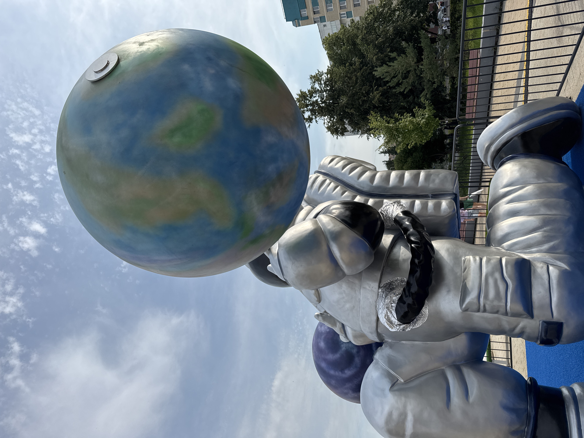 The image depicts a large, metallic sculpture of an astronaut holding a globe of Earth above its head. The astronaut is rendered in a silver, somewhat cartoonish style, with exaggerated features and segmented limbs. The globe is blue and green, representing continents and oceans, and has a small metallic disc on its surface. Behind the sculpture, a purple, swirling spherical object is partially visible, suggesting it might be another planet or celestial body.
The sculpture is situated outdoors in what appears to be a public park or playground area, indicated by a bright blue surface beneath the astronaut and a black metal fence in the foreground. Beyond the fence, a paved walkway and benches are visible, alongside lush green trees and a residential building in the background. The sky is partly cloudy, with a mix of blue sky and white clouds.
This sculpture is located in Zelenograd, Russia, specifically in the vicinity of the "Cosmonauts" monument in Alley of the Cosmonauts. The Alley of the Cosmonauts is a popular public space in Zelenograd, often featuring art installations and memorials related to space exploration.