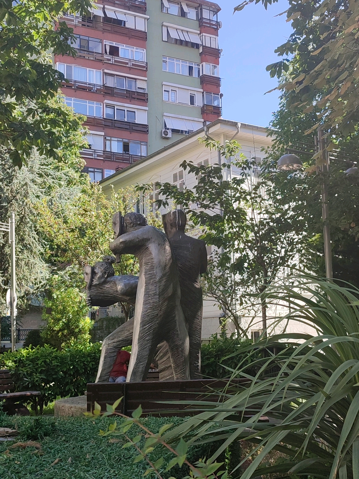 The image depicts a public square in Kadıköy, Turkey, featuring a bronze sculpture of three figures. The sculpture is positioned in the foreground, with lush greenery and trees surrounding it. In the background, a multi-story building with red and green facades dominates the skyline. The building has numerous windows and balconies. A smaller white building is visible in front of the taller structure. The scene is bathed in sunlight, suggesting a clear day. A child wearing a red jacket is partially visible sitting within the sculpture. Benches are placed around the sculpture. The overall atmosphere is tranquil and urban.
