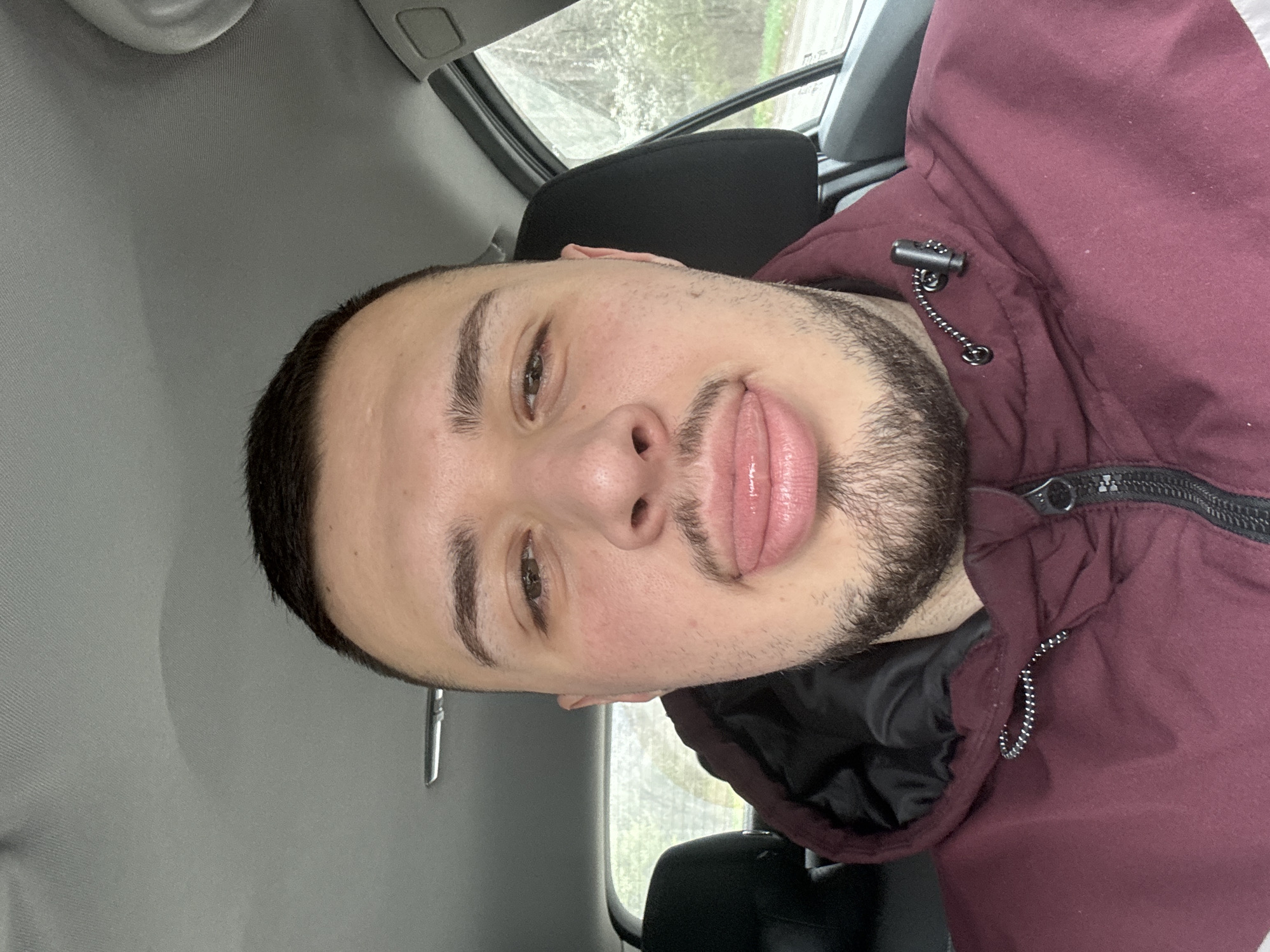 A man is in a car. The man has brown hair, a beard, and is wearing a burgundy hooded jacket. The car's interior is gray. It's daytime, and the trees are visible through the car window. The man is sticking out his tongue. It is not possible to identify the location as Sofia, Bulgaria from the image alone.