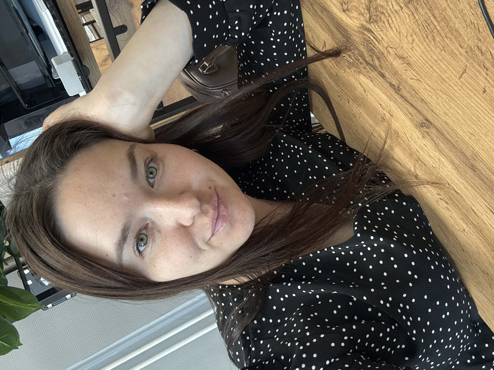 The image shows a woman with brown hair and fair skin, wearing a black and white polka dot dress. She appears to be indoors, possibly in a cafe or office. Her right arm is raised and resting behind her head. A brown purse is visible near her arm. The background includes a wooden surface, a window with blinds, and part of a plant. The location is unclear without further context, but based on the request, it may be Chaikovsky, Russia.