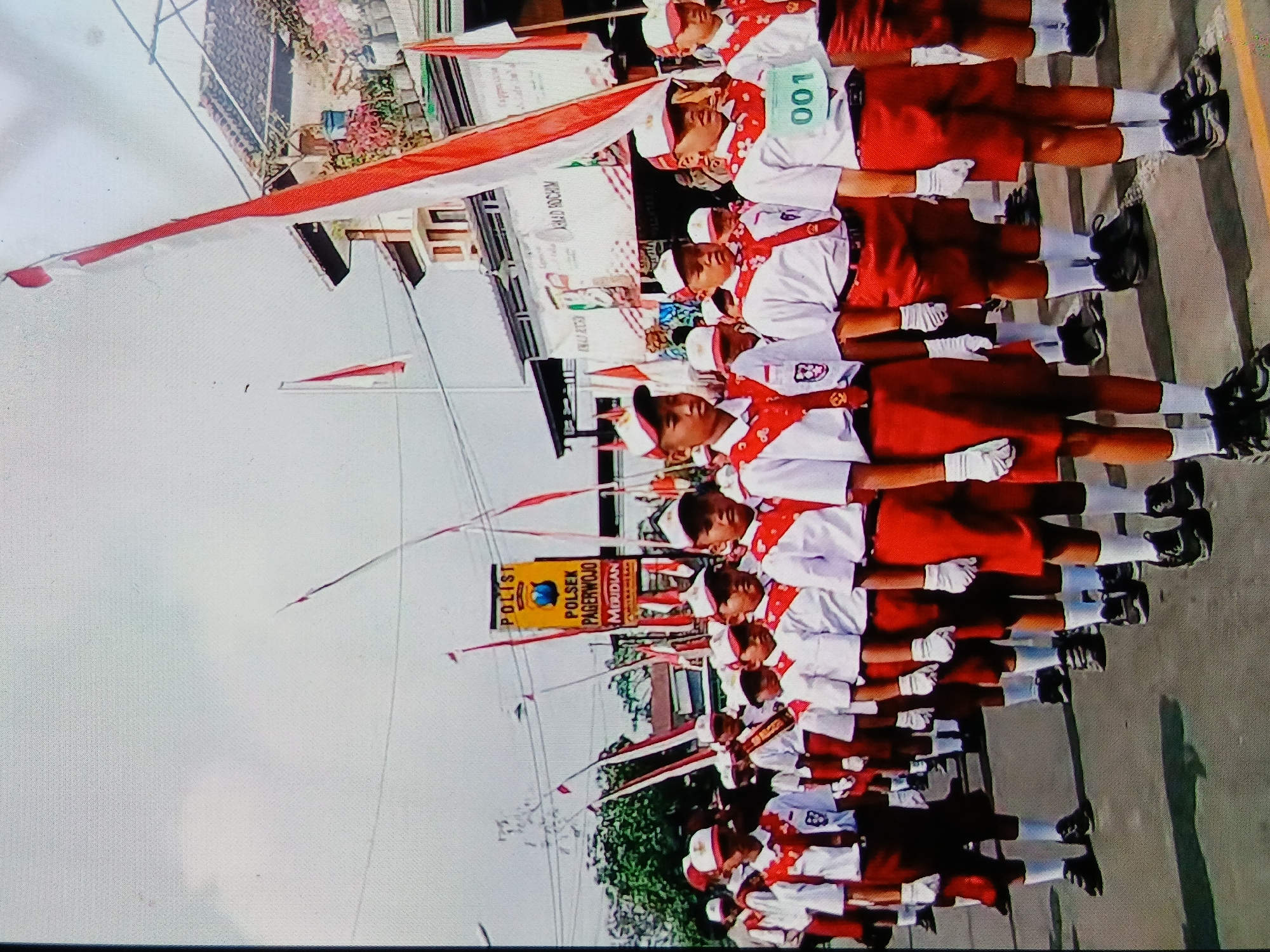 A group of young Indonesian schoolchildren, dressed in white shirts, red shorts, white socks, and black shoes, participate in a parade. They wear red and white sashes around their necks and red and white hats. Some children wear white gloves. Several children have bibs with numbers, including one with "001". The parade appears to be in a street with buildings on the right side. A yellow banner with text, including "POLIST", "POLSEK PAGERWOJO", and "MERIDIAN", is visible. Red and white flags are displayed throughout the scene. The sky is overcast.