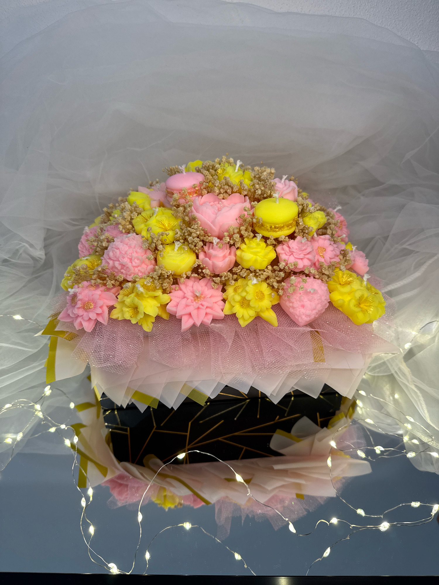 The image displays a decorative bouquet made of candles, arranged in a round, festive presentation. The candles are sculpted to resemble various flowers in shades of pink and yellow, with a few pink macaron-shaped candles interspersed. Small, dried brown floral elements are used as filler. The bouquet is presented in a dark cylindrical container adorned with a geometric gold pattern, and wrapped with layers of white and pink translucent paper and netting. The entire arrangement is placed on a reflective surface, with a white, sheer fabric backdrop. A string of warm white fairy lights is draped around the base of the bouquet and in the foreground, casting a soft glow and creating reflections. The image is well-lit, with the lights from the fairy string contributing to the overall ambiance. There are no people or animals visible in the image. The setting appears to be indoors, possibly a studio or a decorated display area, with no specific architectural details or urban elements indicating a location in Sopot, Bulgaria or anywhere else.