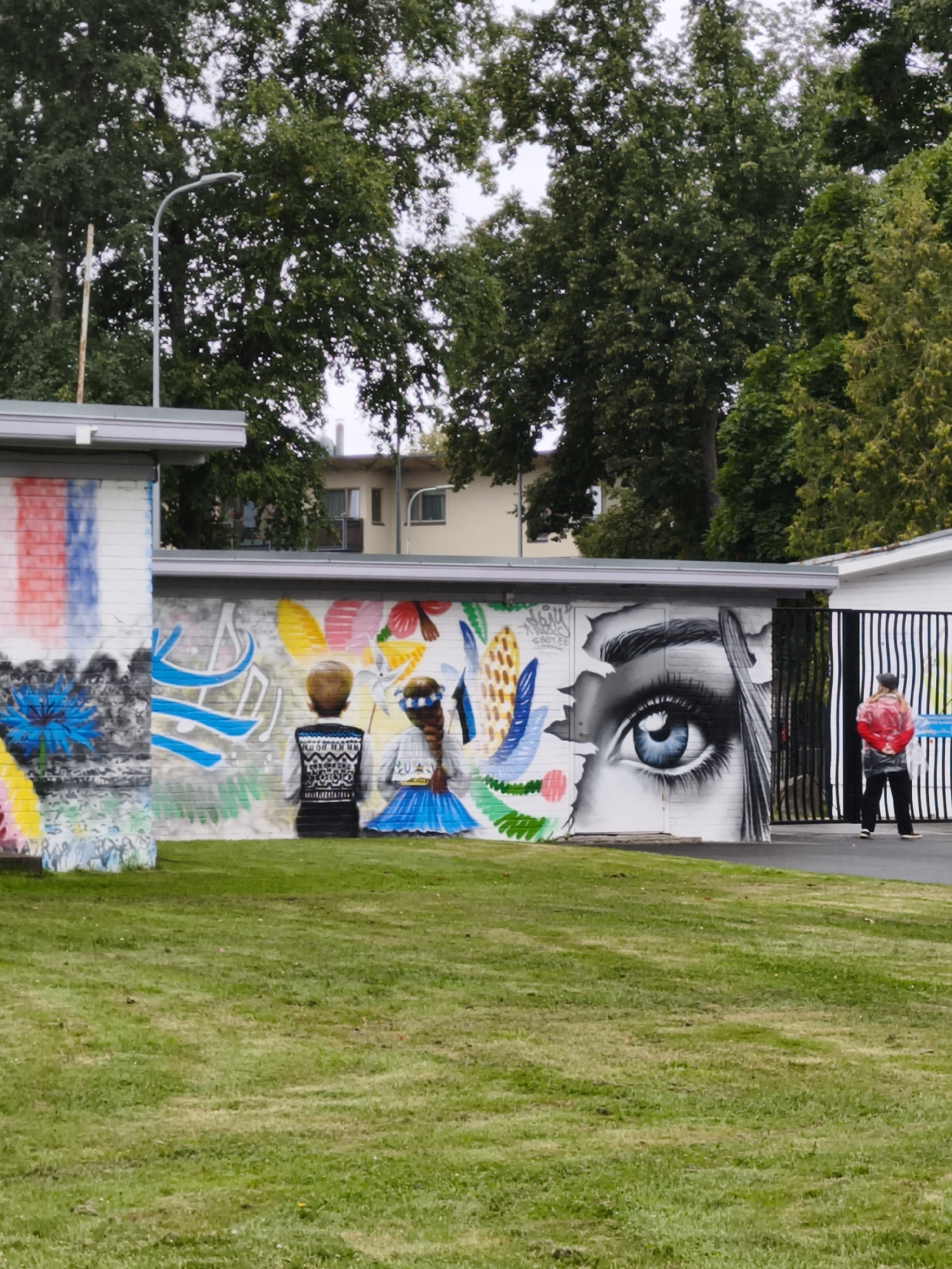 The image depicts a mural on the exterior wall of a building, likely an educational institution given the presence of children depicted in the artwork. The mural features vibrant, colorful designs including abstract shapes, floral motifs, and two figures of children, one boy and one girl. The boy is wearing a patterned sweater, and the girl is in a blue dress and a flower crown. A prominent feature of the mural is a large, realistic eye with a blue iris. To the right of the mural, a black metal fence encloses an area, and a person wearing a red jacket and dark pants stands facing away from the viewer, looking towards the fence. The scene is set outdoors with a grassy lawn in the foreground and trees in the background. The architecture of the building suggests a functional, possibly utilitarian design. The overall impression is one of a public space enlivened by street art. The mural's style and subject matter, particularly the depiction of children and floral elements, along with the potential educational context, might suggest this location to be within a school or community center in Tallinn, Estonia.
