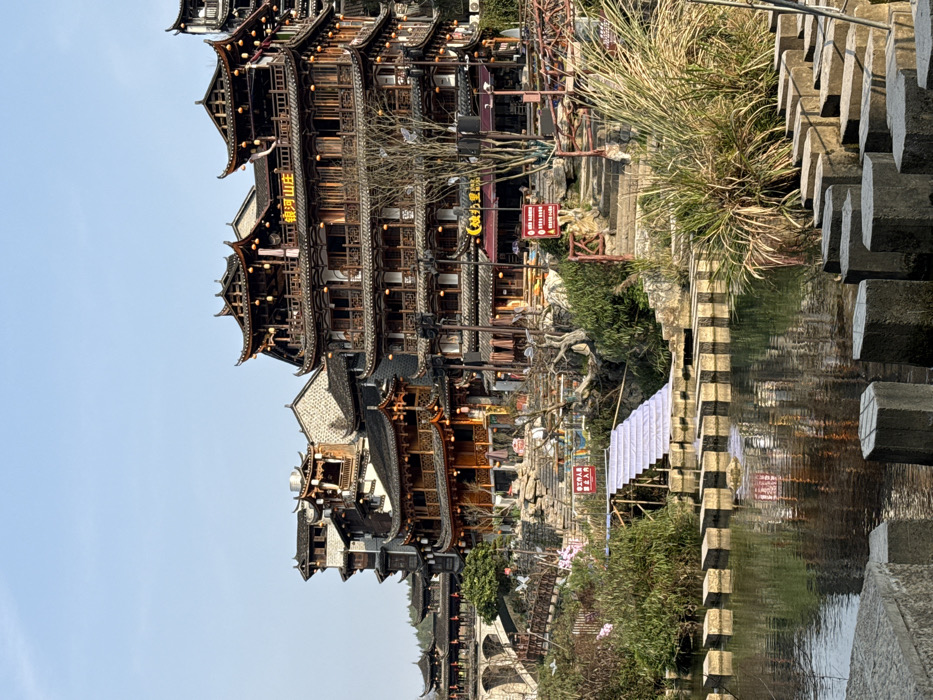 The image depicts a traditional Chinese waterside town in Furong, China, under a clear, bright sky. In the foreground, a series of rectangular and octagonal concrete stepping stones extends across a calm body of water, reflecting the sky and distant structures. Tall grasses and reeds grow along the right bank.
The midground features an intricate arrangement of multi-story wooden buildings with distinctively curved, layered dark tiled roofs and ornate railings, characteristic of traditional Chinese architecture. These structures are built into a hillside, connected by various stone steps and paths. Red lanterns hang from several balconies. One prominent building displays a large yellow sign with Chinese characters reading "银河山庄" (Yín Hé Shān Zhuāng), while another below it has a red sign reading "映月里" (Yìng Yuè Lǐ).
On the left side of the water, a temporary structure, possibly a stage or temporary steps, is draped in white fabric and extends towards the stepping stones. Adjacent to this, a red sign with white Chinese characters is mounted on a wooden frame, indicating a restricted area (likely "工作人员止步" - Staff Only). Further to the left, a stone arch bridge is visible in the distance, spanning across what appears to be another section of the waterway.
The scene includes sparse human presence; distant figures are faintly discernible on pathways and near the water's edge, but their specific actions and identities are not clear. The environment combines natural elements like water, rock, and vegetation with a dense concentration of man-made historical-style buildings, suggesting a scenic and culturally significant location.
