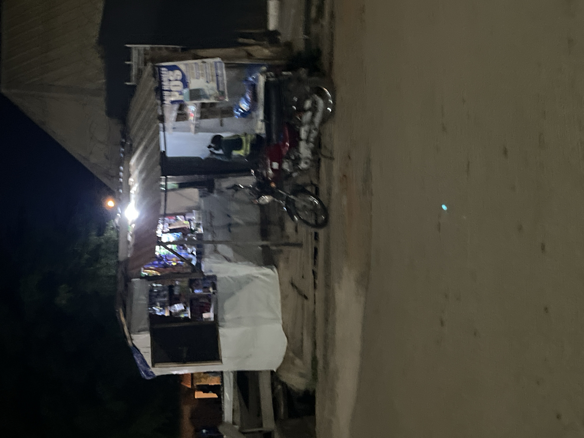 The image shows a night scene in a city, possibly Abuja, Nigeria, featuring a small roadside stall or shop. The stall has a corrugated metal roof and appears to be stocked with various items, including what look like packaged goods and possibly electronics. A red motorcycle is parked in front of the stall. A person wearing a high-visibility vest is visible inside or near the stall. A sign with the text "POS" is prominently displayed on the stall. The surrounding environment is dark, suggesting it is nighttime, and the road in the foreground is paved. The overall impression is one of a typical urban scene at night in a developing city.
