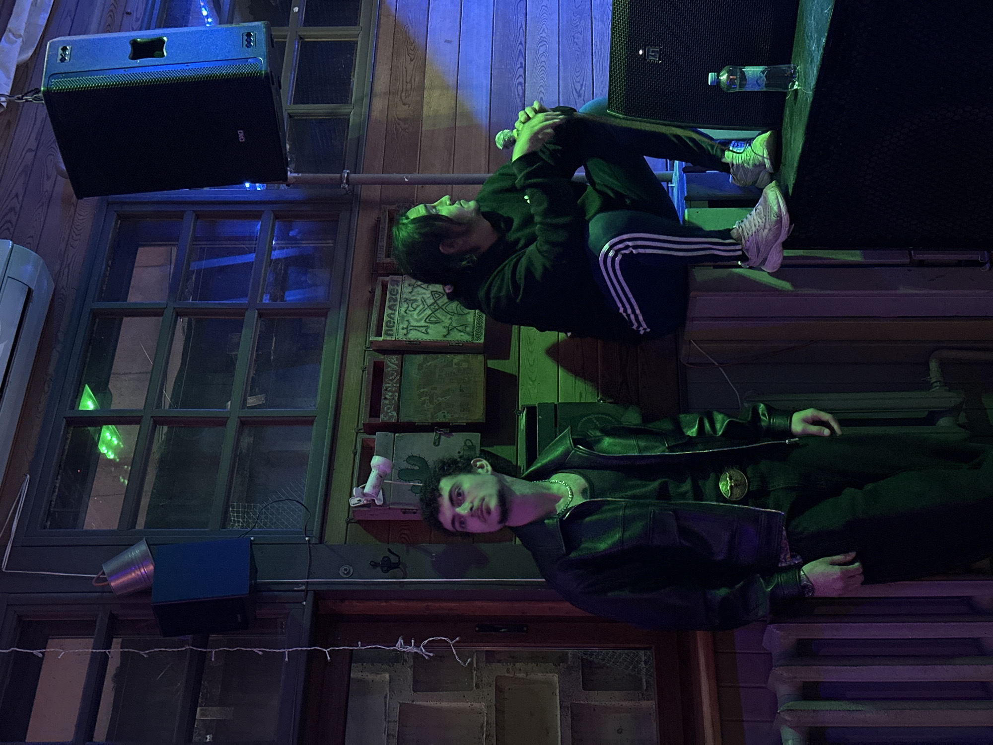 Two people are positioned in a space with a wooden interior and multi-paned windows. One person is sitting cross-legged atop a sound speaker, holding a microphone. They are wearing a dark hoodie and pants with white stripes down the side. Above them is another sound speaker with a water bottle sitting on top. The other person is lying horizontally in front of a door, wearing a dark leather jacket and a silver chain around their neck. The area is lit with blue and green lights, creating a stylized ambiance. A small metal pail is attached to the wall near a window.