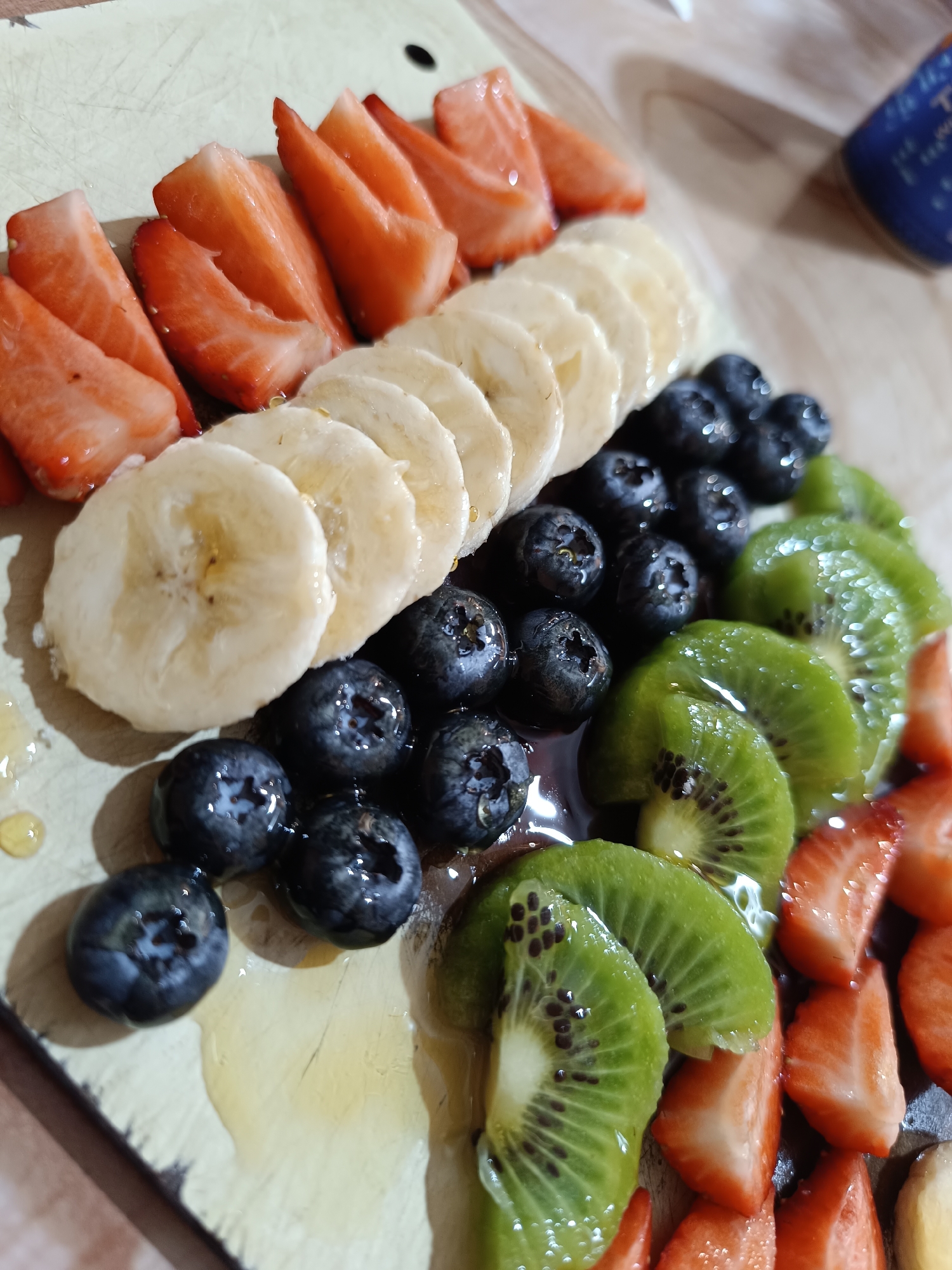 A high-angle shot shows a wooden board arranged with rows of sliced strawberries, bananas, blueberries, and kiwi. The fruit is glistening, suggesting a glaze or syrup. The board is likely indoors under artificial lighting. There is no visible indication of the location being Pleven, Bulgaria. No people are visible.
