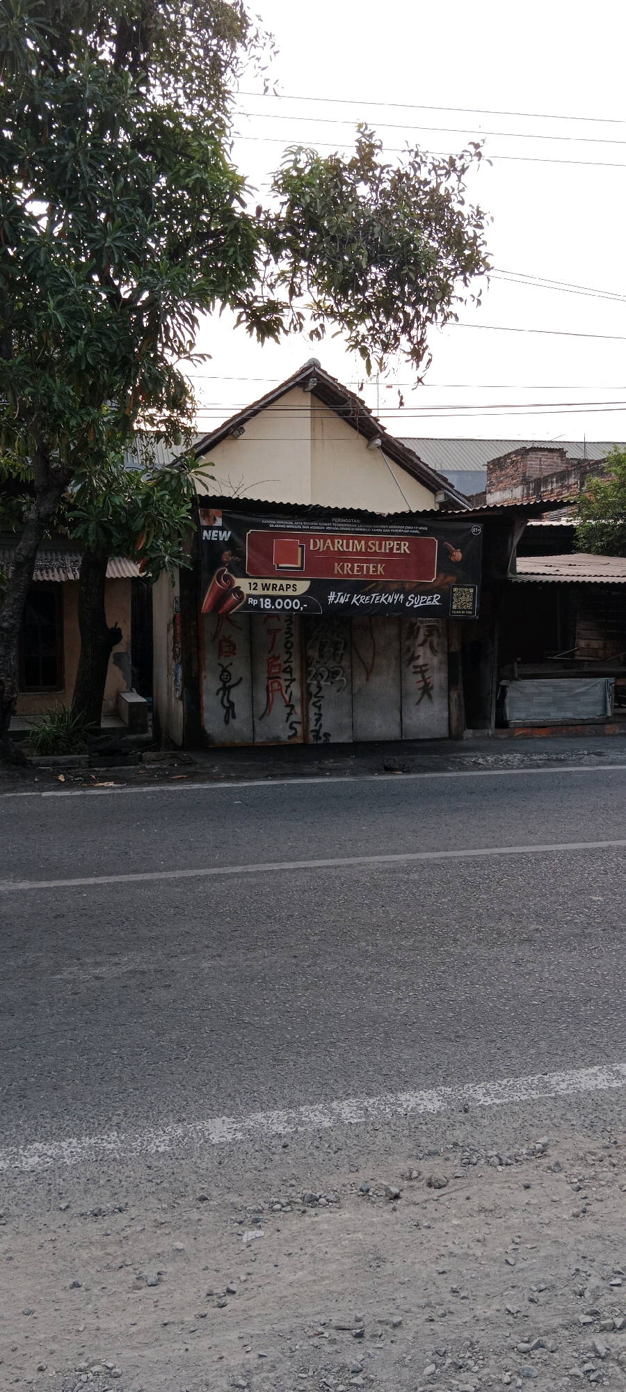 The image captures a street scene in Bojonegoro, Indonesia. In the foreground, a gray asphalt road with a white line is visible. On the right side of the road, a small building with a corrugated metal roof and a banner is situated. The banner displays the brand "DJARUM SUPER KRETEK" along with text in Indonesian. The building's walls are covered with graffiti. To the left of the building, a large tree with green leaves stands. The background shows more buildings and trees under a bright, possibly overcast sky. The overall scene suggests a commercial area or a roadside stall.