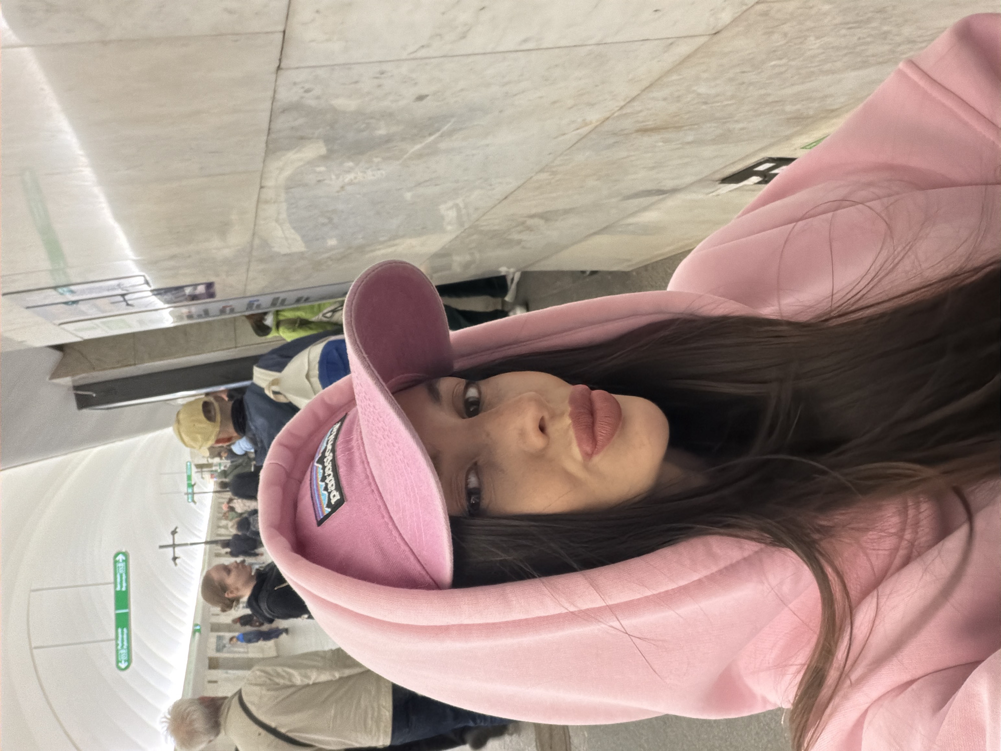 A woman wearing a pink hoodie and a pink Patagonia baseball cap is standing inside Avtovo station in Saint Petersburg, Russia. The station features white marble walls, and there are people walking in the background. Directional signs are visible in the station.