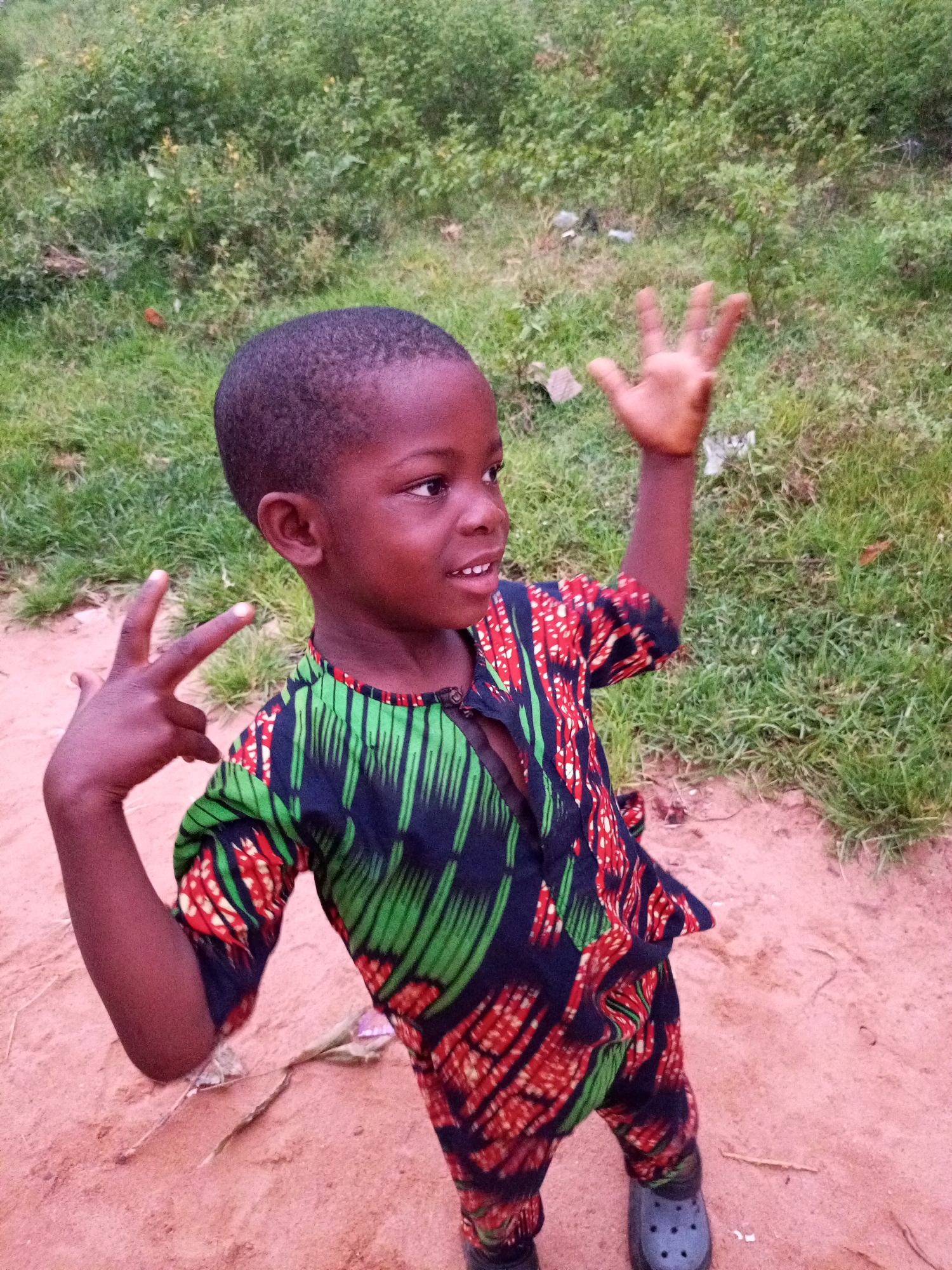 A young Black child, likely male, stands in what appears to be a rural or semi-rural environment. The child is wearing a colorful, patterned African print outfit consisting of a shirt with a mandarin collar and matching pants. The shirt has vertical green and red abstract designs on a dark blue background. The child is looking to the right of the frame, with a slight smile. His right hand is raised with fingers spread, and his left hand is held up, forming a peace sign with his index and middle fingers. The ground beneath the child is reddish-brown dirt, and in the background, there is a patch of green vegetation. The lighting suggests it is daytime, possibly overcast. The setting is consistent with an outdoor scene in a village or town in Nigeria, such as Tempure.