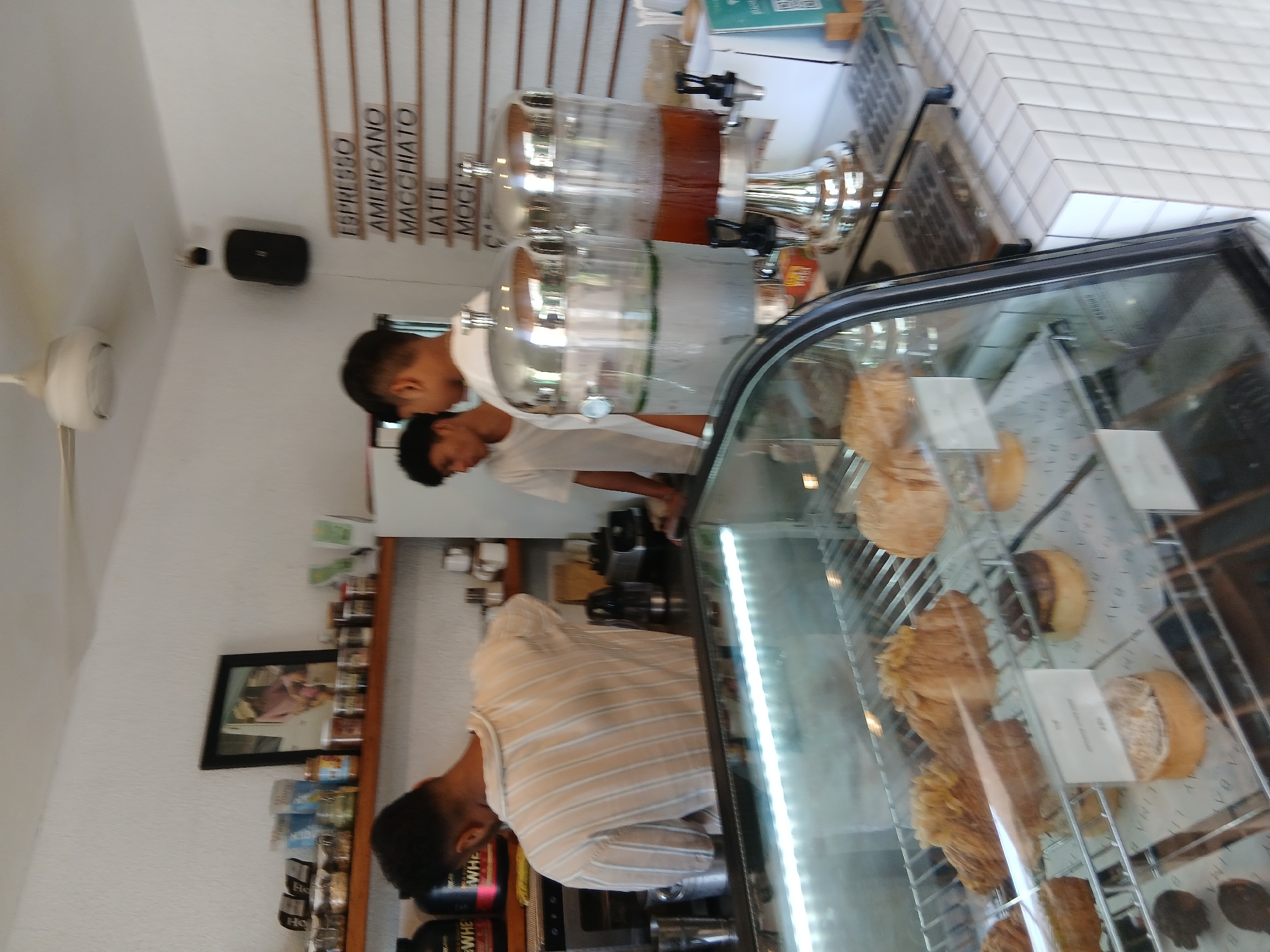 This image captures the interior of a cafe in North Kuta, Indonesia, likely a small business based on its setup. In the foreground, a refrigerated display case exhibits various pastries and baked goods, including croissants and circular tarts. Behind the counter, two young men, presumably staff, are visible. One, wearing a light-colored, striped shirt, appears to be engaged in preparing a drink behind a coffee machine. The other, in a white t-shirt, stands to his right, observing. To the right of the men are two large, silver beverage dispensers, one filled with a clear liquid and the other with a darker, amber-colored liquid, likely iced tea or juice. Above and behind them, shelves display various items, including jars, bottles, and a framed picture. A menu board with a list of coffee drinks like "ESPRESSO," "AMERICANO," "MACCHIATO," "LATTE," and "MOCHA" is mounted on the wall to the right, with a black speaker mounted higher up. The cafe features white tiled walls and a ceiling fan. The overall impression is a functional and inviting space for a local coffee shop.