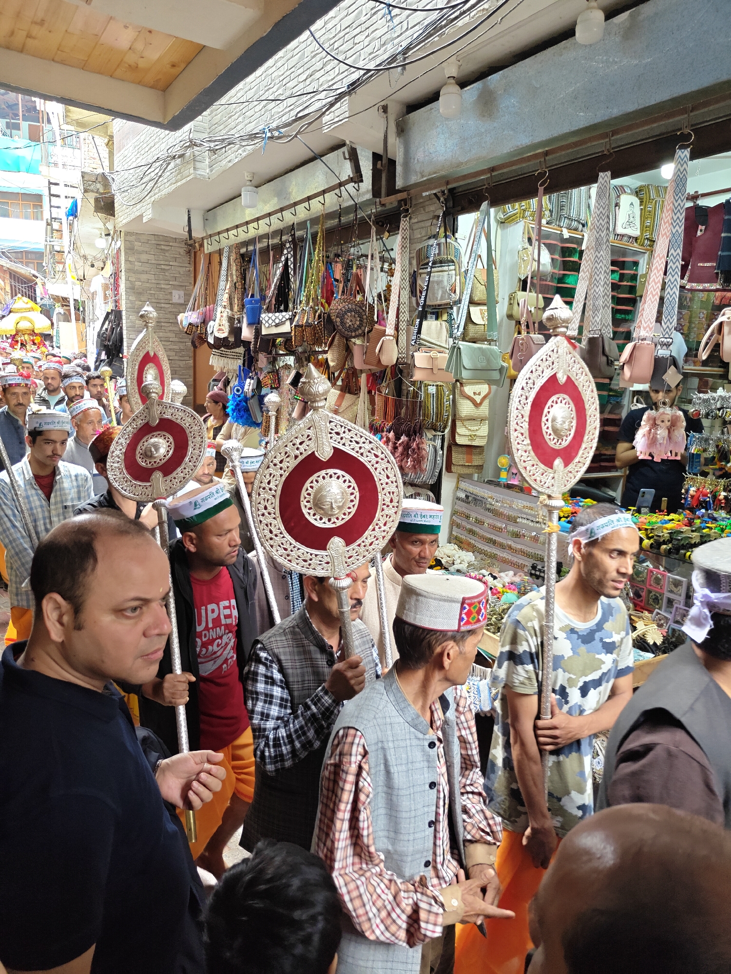 This image captures a vibrant procession of men moving through a narrow, bustling street in Kasol, India. The scene appears to be a daytime event, possibly a religious or cultural parade, given the traditional attire and ceremonial objects.
The main subjects are numerous men, many of whom are wearing distinctive Himachali caps, which are round and often feature colorful patterned bands. Several participants are holding tall, ornate ceremonial staffs, known as 'chharis,' that have large, decorative circular or oval tops made of silver-toned metal with red inlays and a central facial motif or symbol. Some men are dressed in traditional Indian garments, including saffron/orange lower wraps (dhotis) and checked shirts with waistcoats, while others wear more casual modern attire like t-shirts.
The street is lined with various shops, whose merchandise spills out into the walkway. Prominently displayed are numerous handbags, slings, and wallets, hanging from racks and shelves. Other visible items in the shops include dolls, toys, and small trinkets. The buildings flanking the street are multi-storied with diverse architectural finishes, and a network of electrical wires is visible overhead.
Visible text on some Himachali caps and a bandana includes "जय बजरंग बली हनुमान जी" (Jai Bajrang Bali Hanuman Ji) and "प्रजापति श्री हेम" (Prajapati Shri Hem), suggesting a devotion to Lord Hanuman. A red t-shirt has partially visible text, possibly "SUPER DRY," and a handbag strap reads "FASHION FOREVER." The participants appear focused and composed as they move along the crowded lane, creating a dynamic snapshot of local culture and commerce.