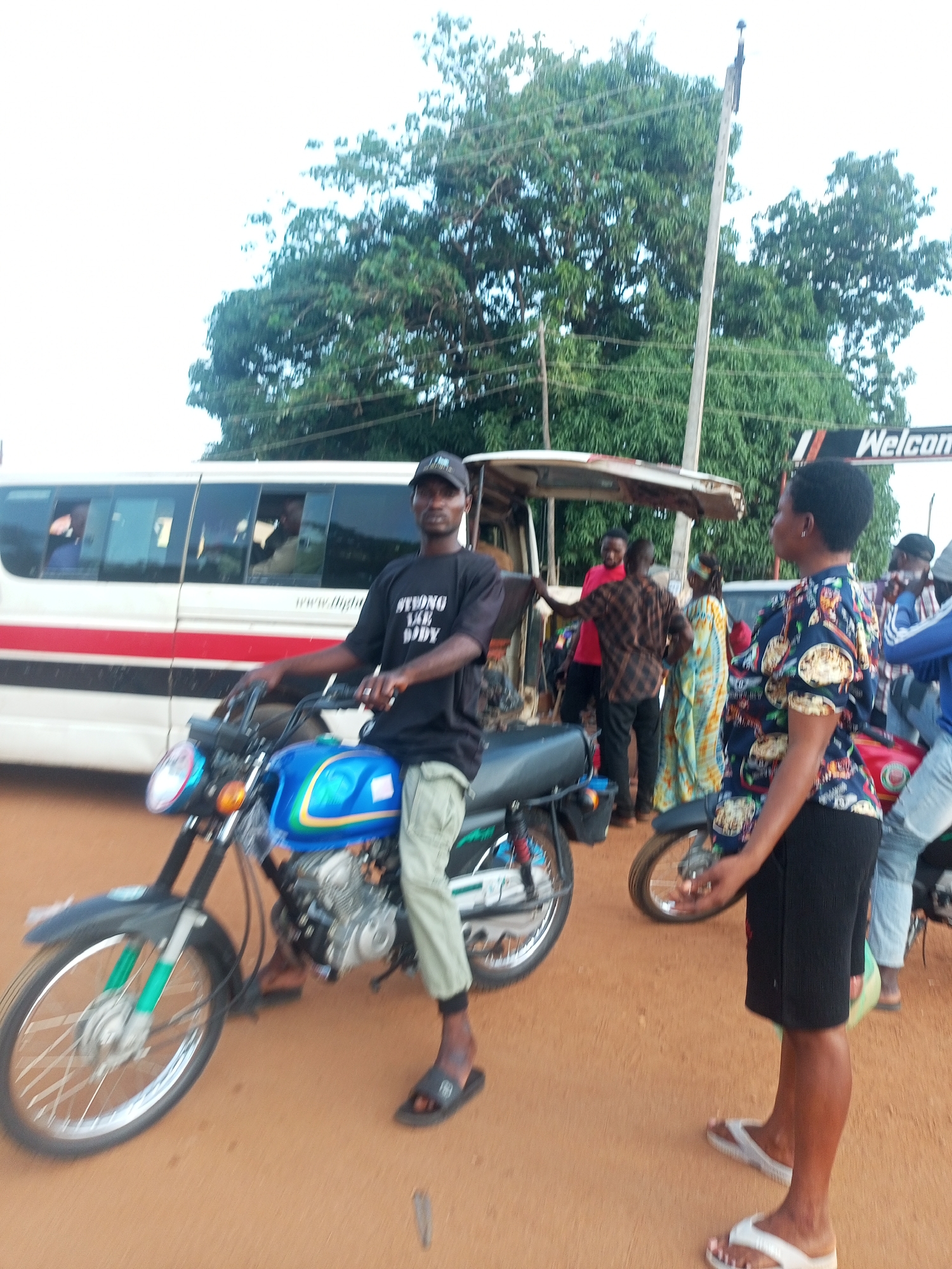 The image depicts a street scene in an unknown city. A man wearing a black t-shirt with white writing and a baseball cap is sitting on a blue motorcycle. He is wearing light-colored pants and sandals. Standing beside him is a woman wearing a patterned blouse and black shorts, also wearing sandals. In the background are several other people, a bus, and a sign that reads "Welcome". The ground is a reddish-brown color. Overhanging foliage from trees are visible in the background.