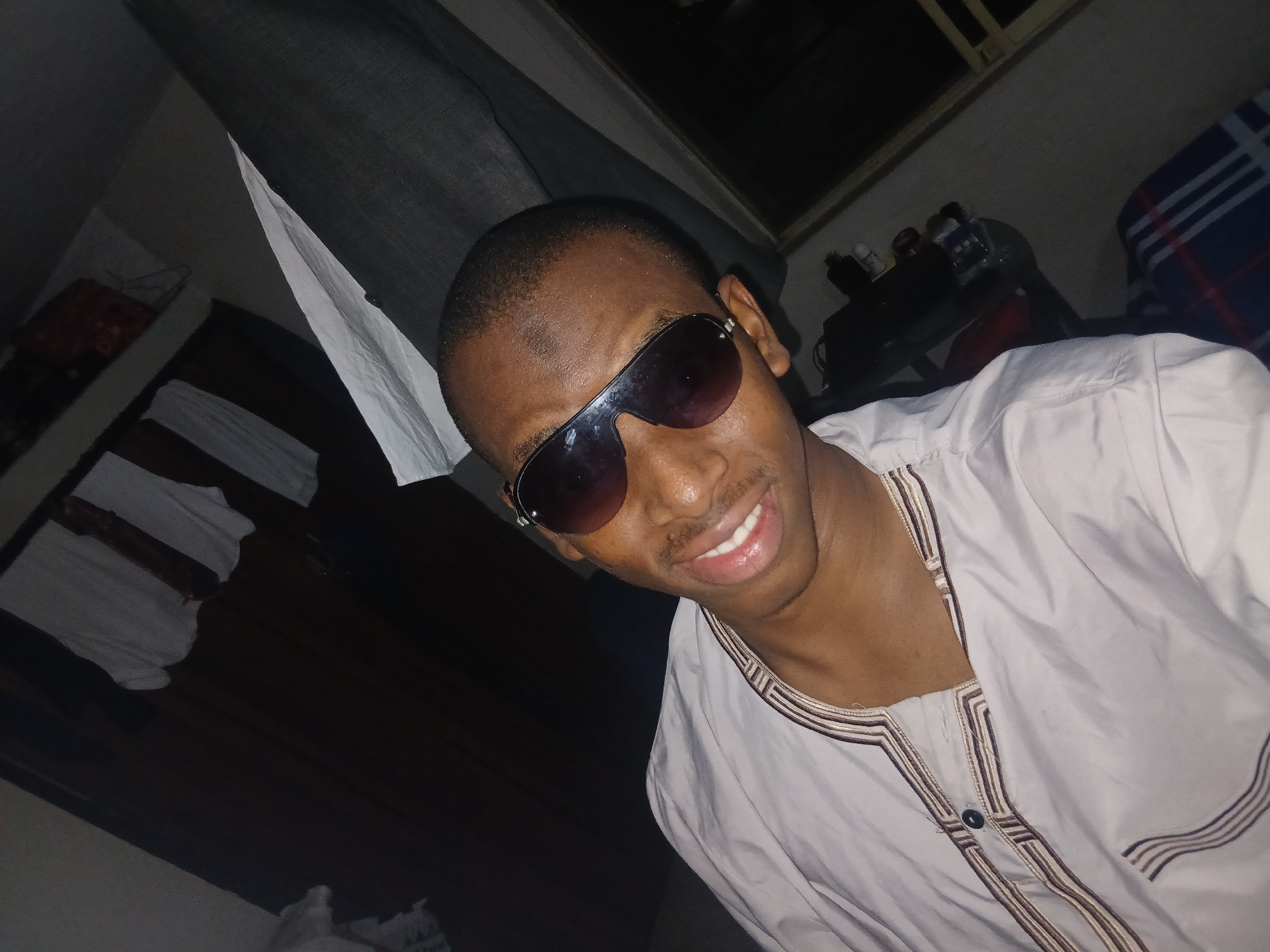 A young Black male wearing sunglasses and a light-colored traditional garment with ornate brown and beige embroidery is captured in a close-up, self-taken photograph, likely in a bedroom in Lagos, Nigeria. The individual's head is tilted, and he is smiling, with his teeth visible. The background shows a dark wooden wardrobe with white garments hanging inside, and a large piece of dark grey fabric, possibly a curtain or clothing draped over furniture, dominates the right side of the frame. A dark table or shelf with several bottles and objects is visible behind the subject. The overall lighting is dim, suggesting an indoor setting at night.