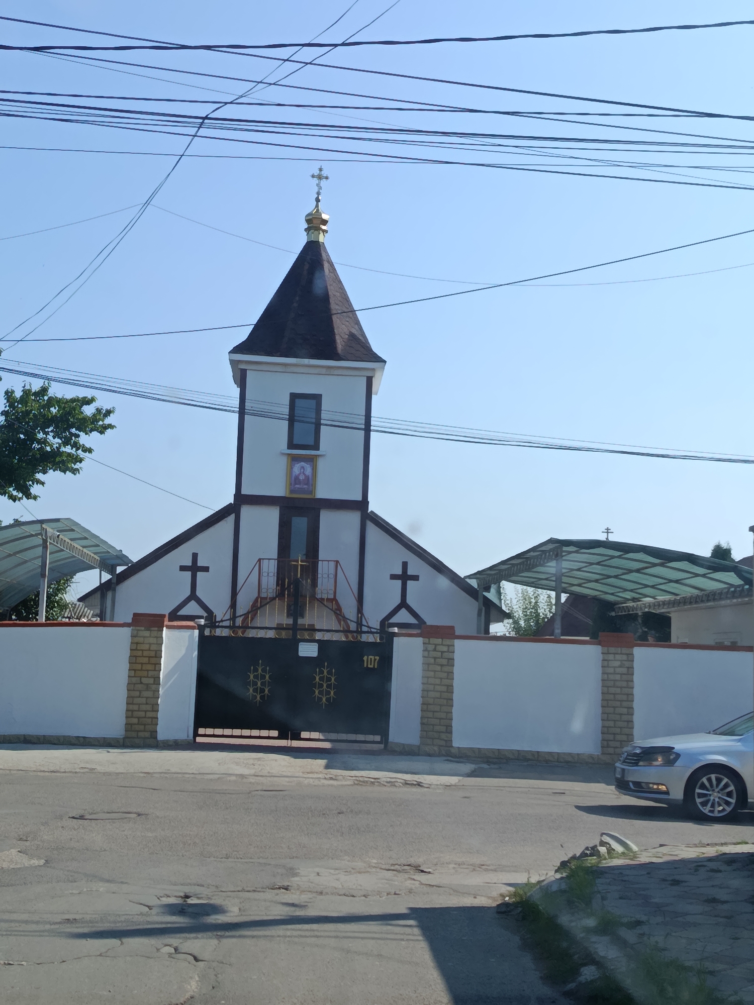 The image shows a church building with a steeple topped with a cross, located in the city of Chișinău, Moldova. The church has a white facade with dark trim and a dark-colored roof. There is a gated entrance to the church property, with the number "107" visible on the gate. A silver car is parked to the right of the frame, and overhead power lines crisscross the sky. The environment appears to be a street scene with a paved road and sidewalks. There are no people visible in the image, and the overall impression is one of a religious structure within a residential or mixed-use urban area.