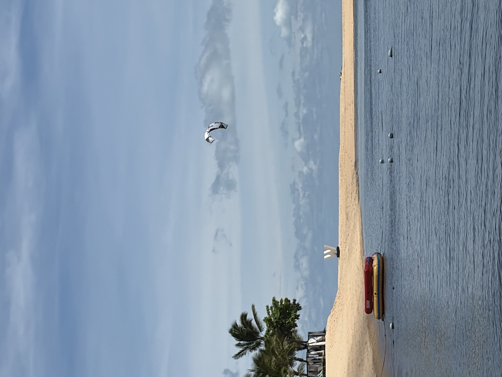 The image captures a serene beach scene in Baan Bo Phut, Thailand. A calm body of blue water occupies the foreground, with several white buoys dotting its surface. In the middle ground, a sandy beach is visible, backed by a line of palm trees and some resort structures. To the left of the beach, a red and yellow inflatable raft is anchored in the water. Further inland on the beach, a large white "W" shaped sculpture stands prominently. In the sky, a kiteboarder is captured mid-air, with a distinctive black and white kite. The sky is a pale blue, with scattered clouds. The overall atmosphere is one of leisure and outdoor activity.