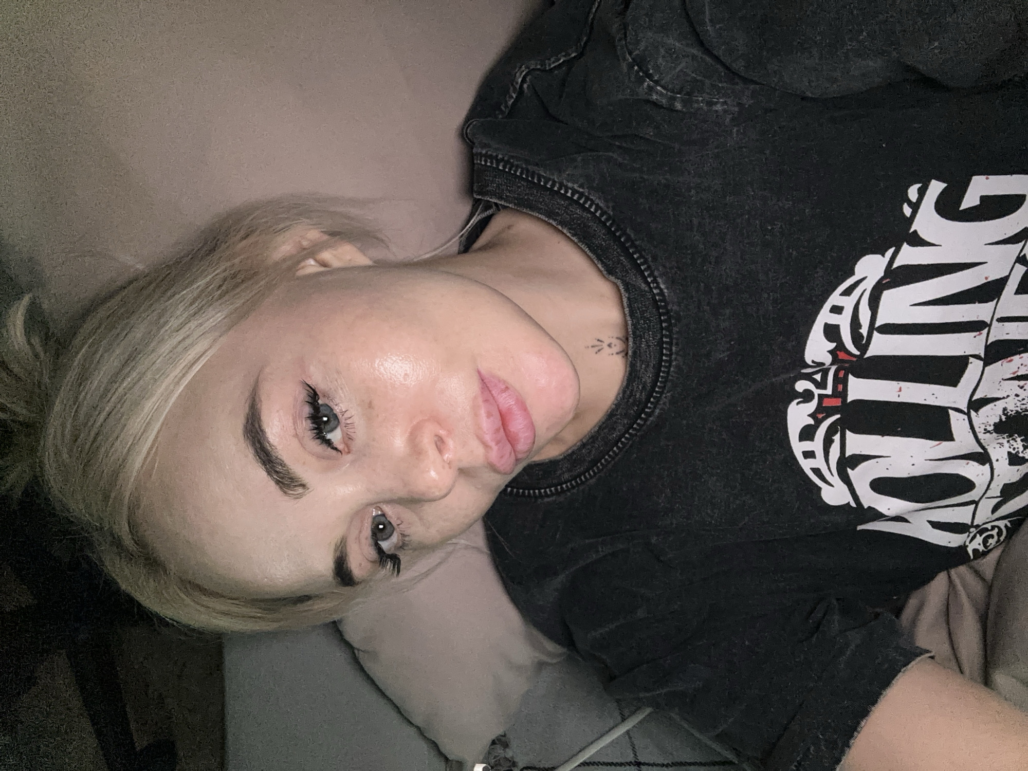 The image shows a fair-skinned person with blonde hair pulled back into a loose bun. They are lying down, possibly on a bed or couch, with their head resting on a pillow. They have light-colored eyes and are wearing false eyelashes. The person is wearing a dark-colored t-shirt with a white graphic design on the front. A small tattoo is visible on their neck. The background is out of focus, showing a plain wall and bedding. The image does not contain information to conclude it was taken in the city of Высокий, Russia.
