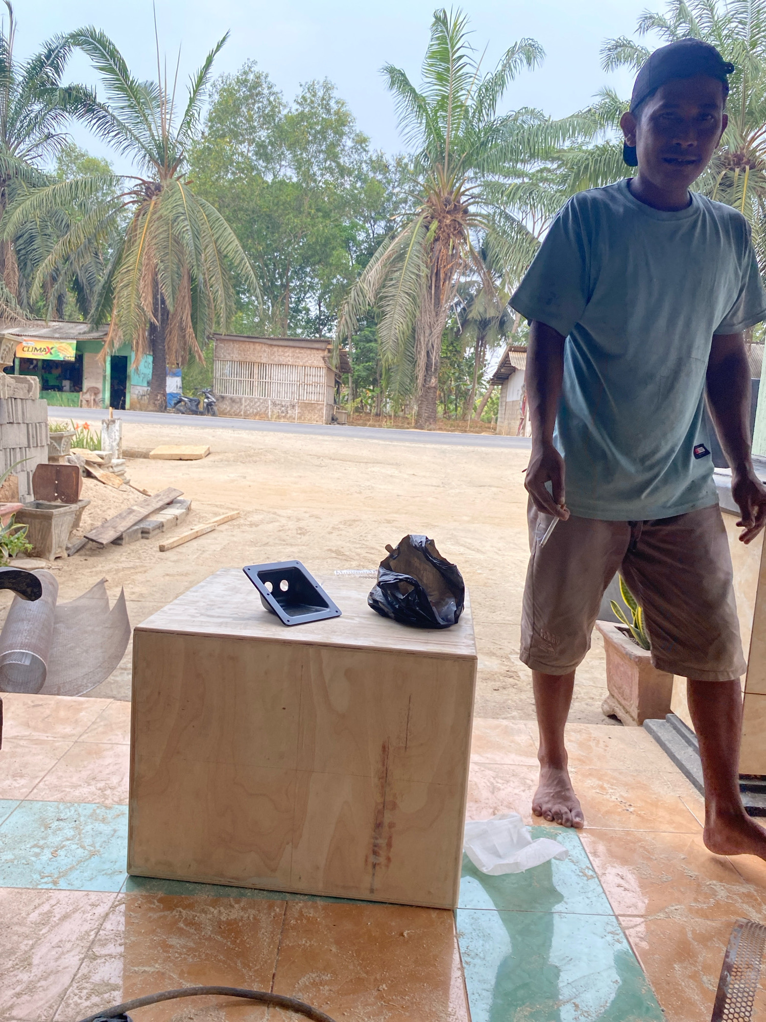 A man wearing a light blue t-shirt and brown shorts stands on a tiled porch, holding a cigarette in his right hand. He is positioned to the right of a large, unfinished wooden box. In front of the box, on the left side, there is a black plastic handle with two openings. To the right of the handle, on the box, is a crumpled black plastic bag. The background shows a dirt road with palm trees and a few simple buildings. The scene appears to be in a rural area of Mojo Pahit, Indonesia.