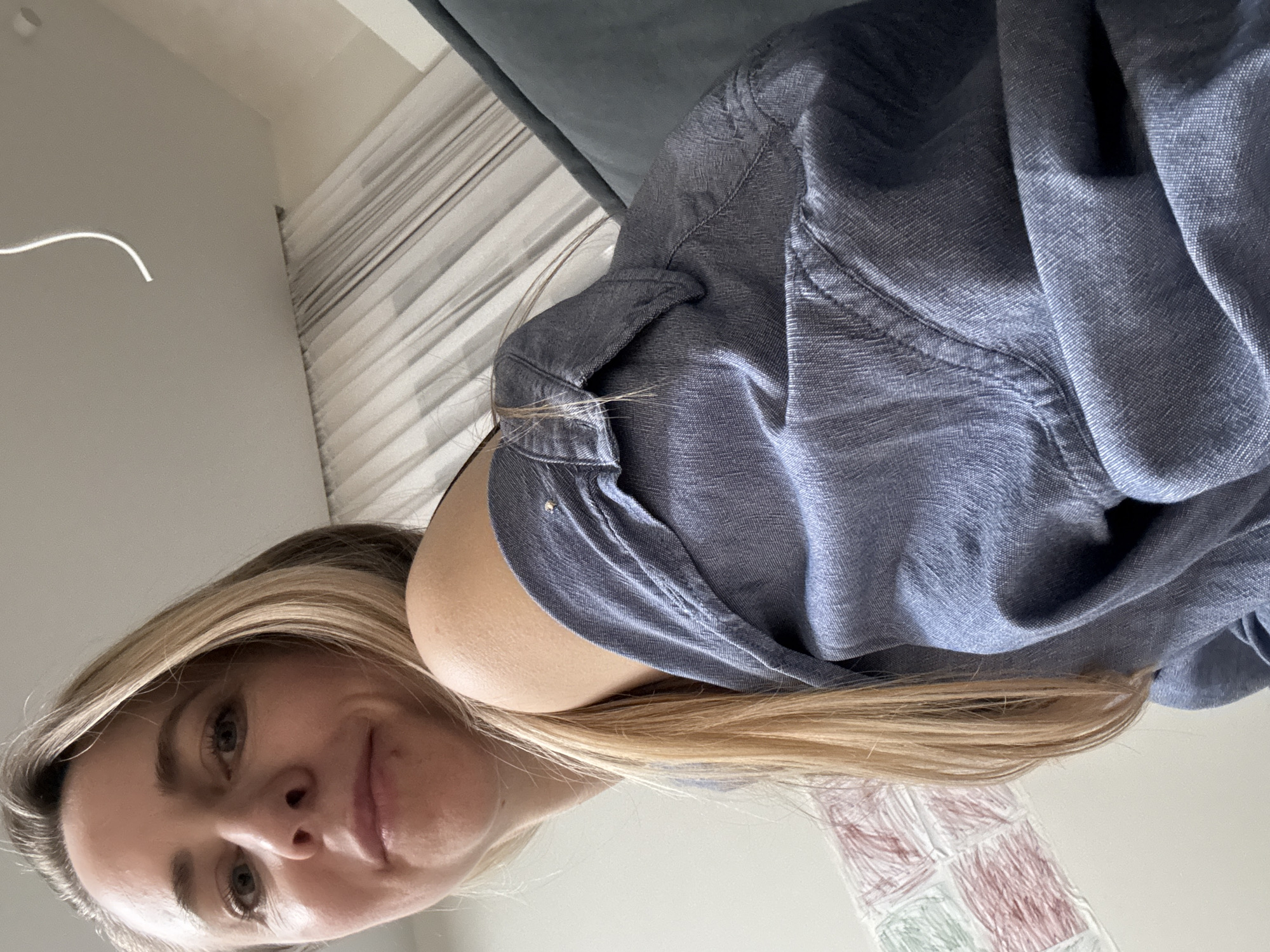 The image shows a woman with blonde hair wearing a blue shirt. She is looking at the camera. In the background, there is a window with blinds and a gray sofa. The location cannot be determined from the image.