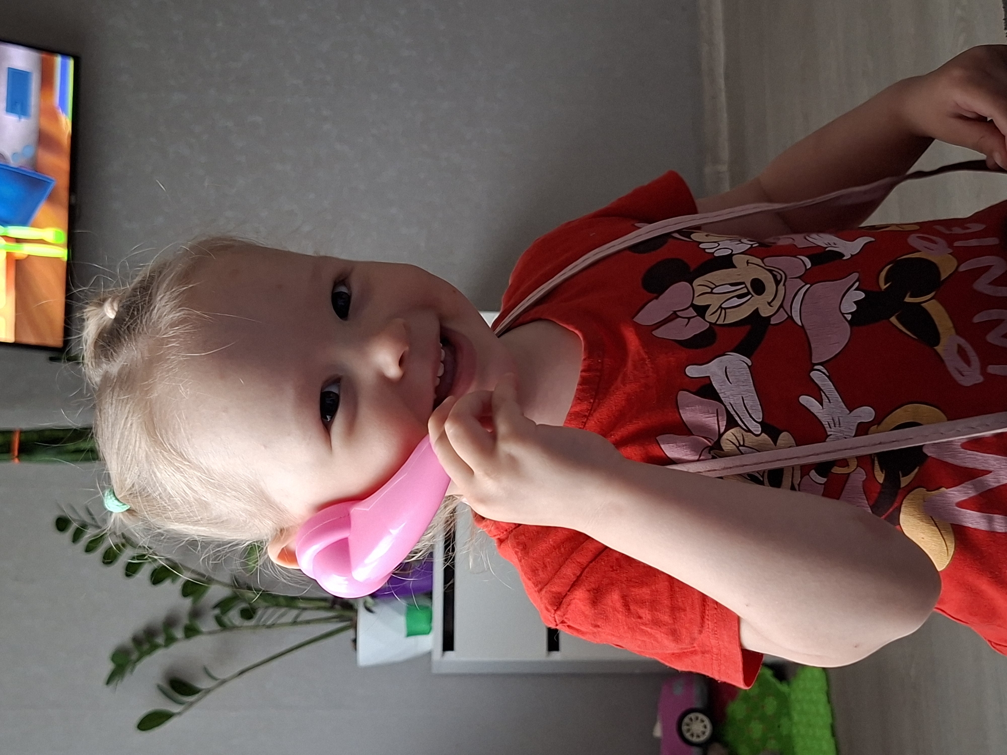 A young, light-skinned child with blonde hair is holding a pink toy phone to their ear, smiling at the camera. They are wearing a red t-shirt with cartoon characters printed on it and a light pink shoulder strap, possibly from a bag or purse. The child is indoors, as evidenced by a television screen showing a cartoon in the background, as well as a potted plant and a piece of furniture. The location in Karsha, Russia, is not possible to discern from the given image.