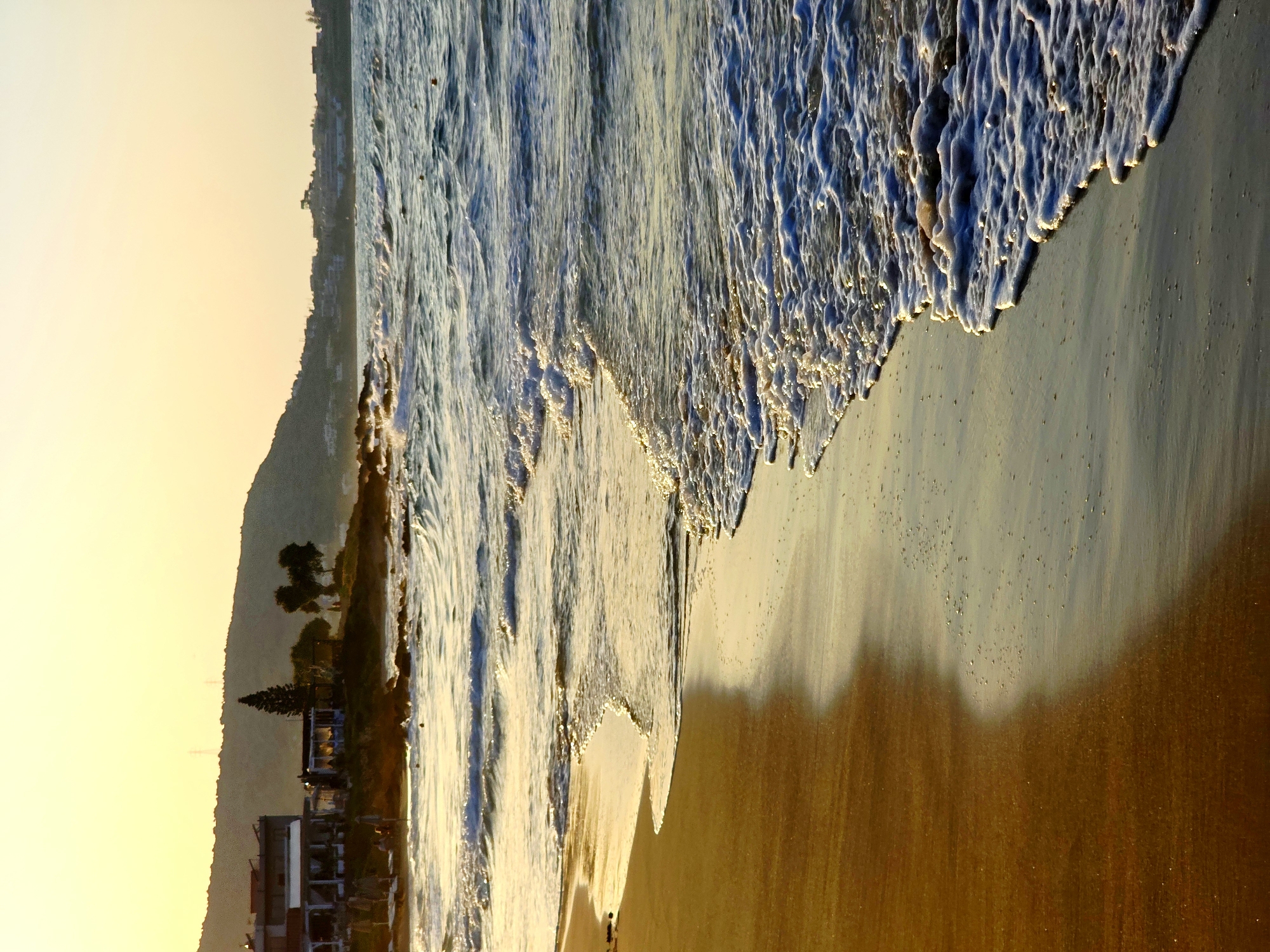 The image depicts a beach scene at sunset in Malia Municipal Unit, Greece. The foreground is dominated by the wet sand of the beach, reflecting the warm hues of the setting sun. Gentle waves are seen lapping at the shore, creating a foamy edge. In the mid-ground, the sea stretches out, with some waves and a few buoys visible.
In the background, a rocky coastline is evident, with a few buildings situated on the hillside overlooking the sea. These buildings appear to be a restaurant or a complex of shops and cafes, with outdoor seating visible. A few trees, including a tall conifer, are scattered amongst the buildings. Further in the distance, rolling hills and more distant buildings suggest the presence of a town or village along the coast. The sky is a soft gradient of yellow and orange, characteristic of a sunset. There are no identifiable people in the foreground or mid-ground; any figures on the distant buildings are too small to discern clearly. The overall atmosphere is tranquil and serene, with the warm light of sunset casting a golden glow over the landscape.