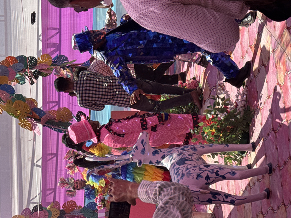 The scene depicts an outdoor event or celebration taking place under a white canopy with pink fabric draping, located in Malasar, India. The ground is covered in pink and light pink patterned tiles or mats, with areas of planted red and green foliage. Overhead, a decorative arch of colorful, leaf-shaped cutouts in pink, yellow, blue, and orange is visible.
In the immediate foreground, to the left, a person's arm and part of their torso are visible, wearing a light-colored, possibly patterned, long-sleeved shirt, with their left hand holding a black smartphone. Adjacent to them, a white statue of a deer or fawn with blue floral and botanical patterns stands upright, facing right.
In the mid-ground, a person wearing a bright pink, sequined or reflective suit with a matching top hat and ruffled collar/cuffs stands facing right. To their right, a person in a dark plaid shirt and dark pants is seen from the back, partially obscured. Further to the right, a person dressed in an iridescent blue, geometric-patterned suit with a matching top hat is in motion, appearing to dance or step forward, with their right arm extended. Part of a person in a light pink textured or polka-dotted shirt is visible in the far foreground right.
In the background, under the canopy, a person wearing elaborate, colorful headwear and a multi-colored costume is visible behind a table adorned with flowers. Other individuals are present in the background, some partially obscured. Bright sunlight casts distinct shadows on the ground.