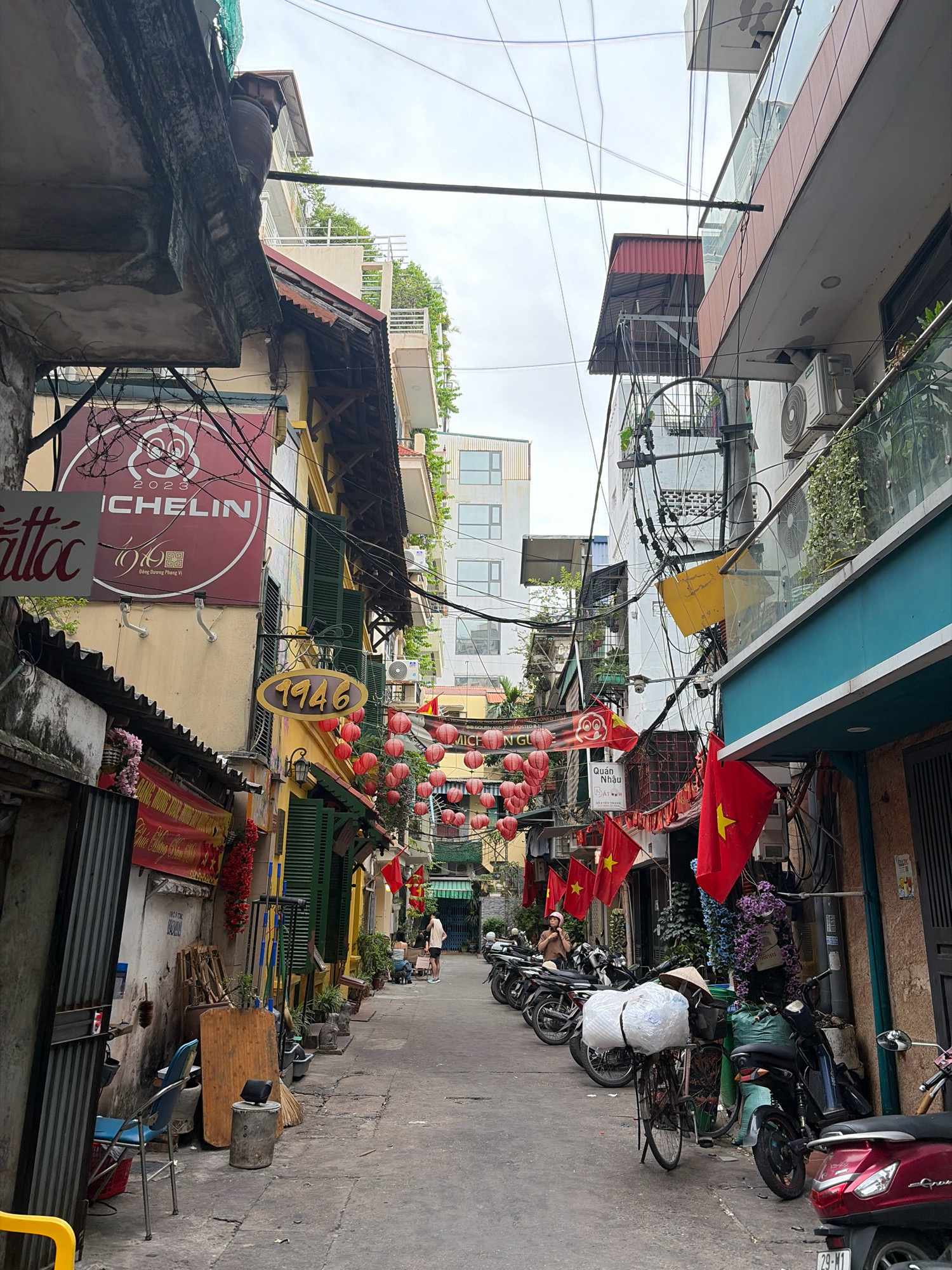 The image depicts a narrow, overcast street in Hanoi, Vietnam, likely in the Old Quarter, characterized by dense urban architecture and a mix of residential and commercial establishments. The street is lined with multi-story buildings, some appearing older with weathered facades and exposed wooden beams, while others are more modern with concrete and glass. A prominent sign reads "2023 MICHELIN," suggesting a connection to culinary recognition. Red Vietnamese flags are strung across the street and displayed on buildings, along with numerous red lanterns.
Several motorbikes are parked along the right side of the street, indicating a common mode of transportation. A person wearing a light-colored jacket and a helmet is seen near the parked motorbikes, possibly about to ride. Further down the street, another person is walking away from the camera. Various items are stored along the sides of the buildings, including wooden planks, furniture, and potted plants. One building on the right has a turquoise-colored facade element. The sky is cloudy and overcast. The scene conveys a sense of everyday urban life in a bustling Vietnamese city.